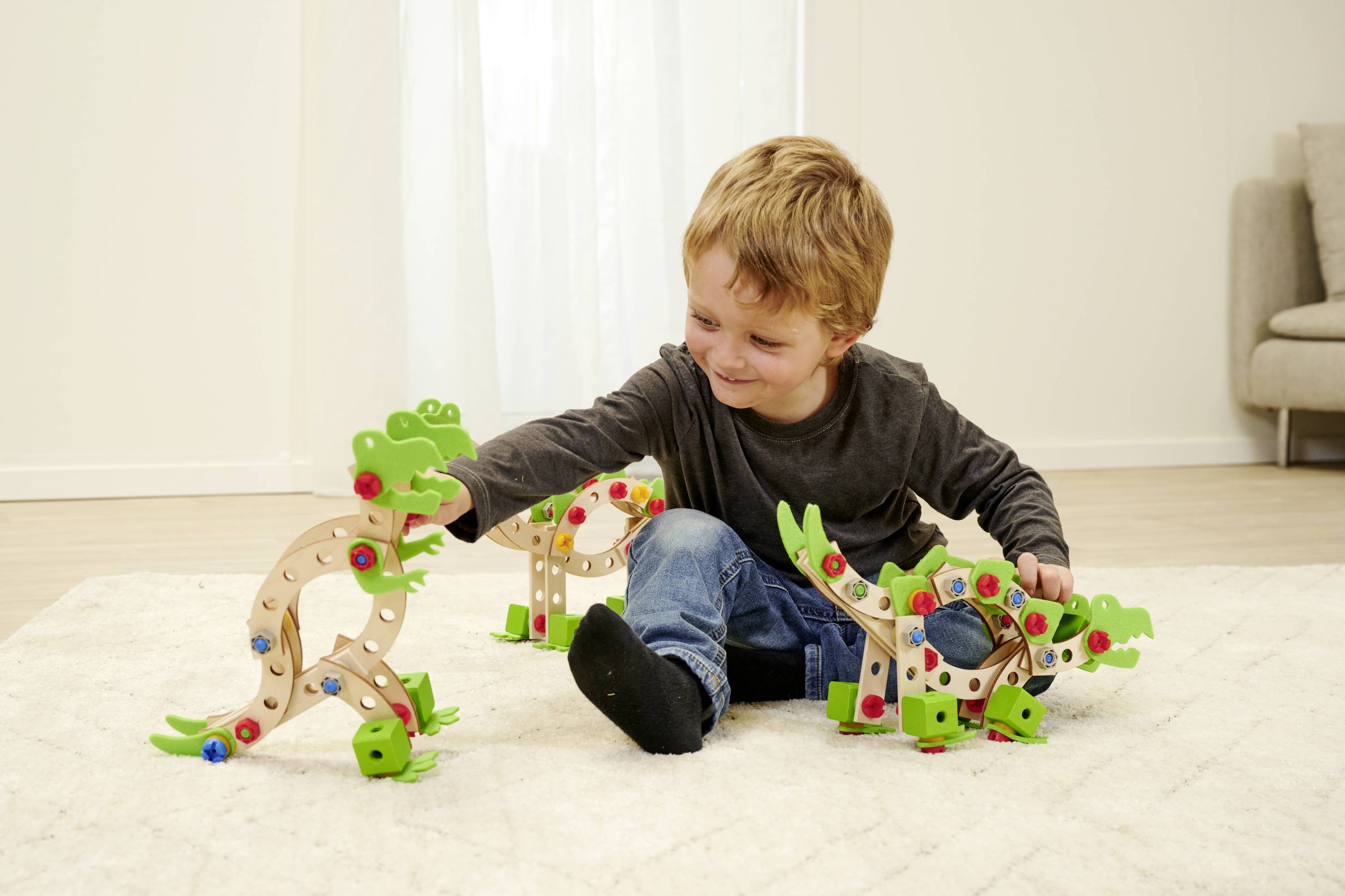 Een kind zit op een lichte vloerkleed en speelt met houten blokken in de vorm van dinosaurussen. Op de achtergrond staat een bank.
