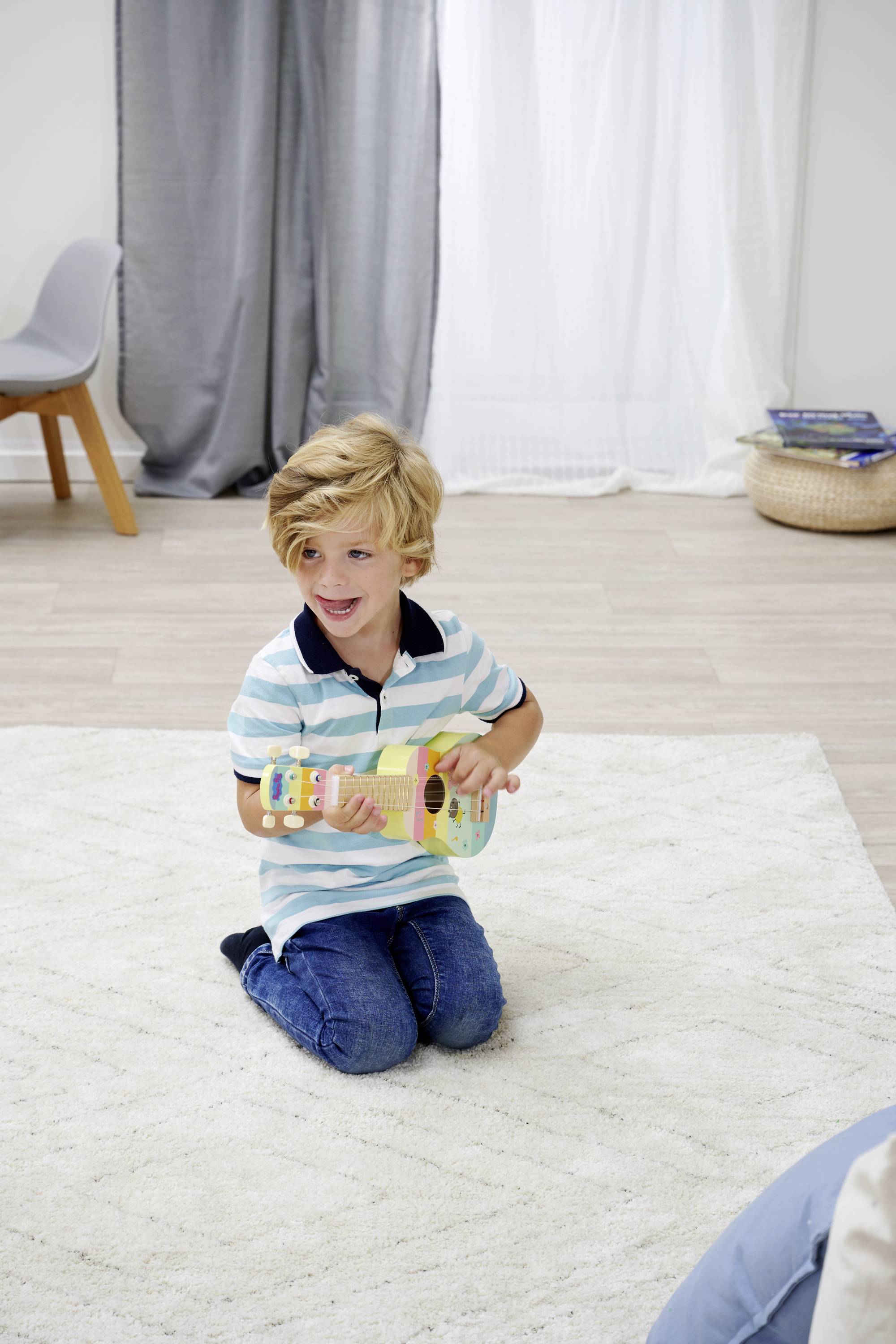 Een jongen zit op een vloerkleed en speelt met een kleine speelgoedgitaar. Op de achtergrond zijn een stoel en gordijnen te zien.