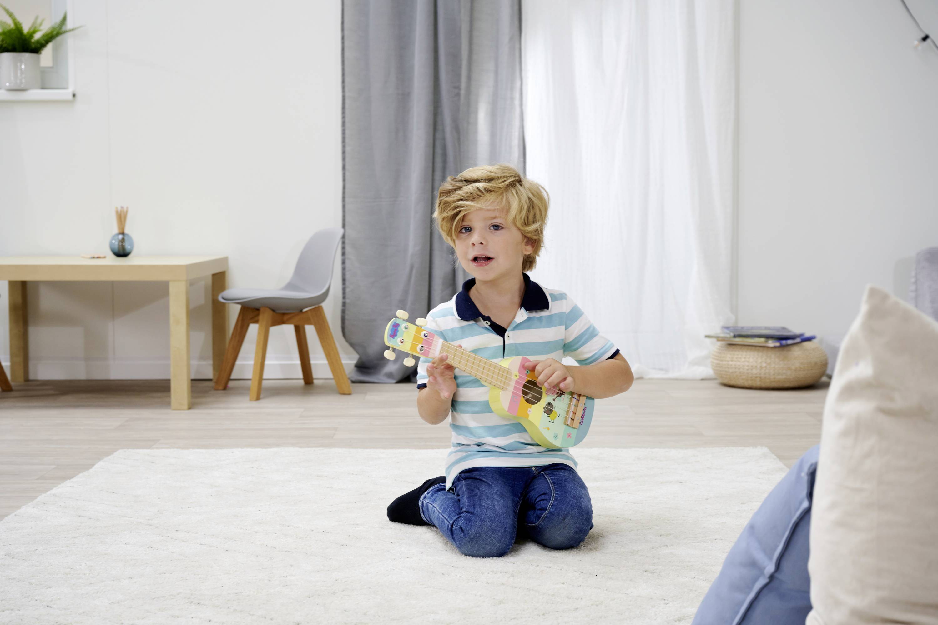 Een kind zit in de woonkamer op een vloerkleed en speelt op een kleurrijke ukelele. Op de achtergrond zijn meubels en gordijnen zichtbaar.