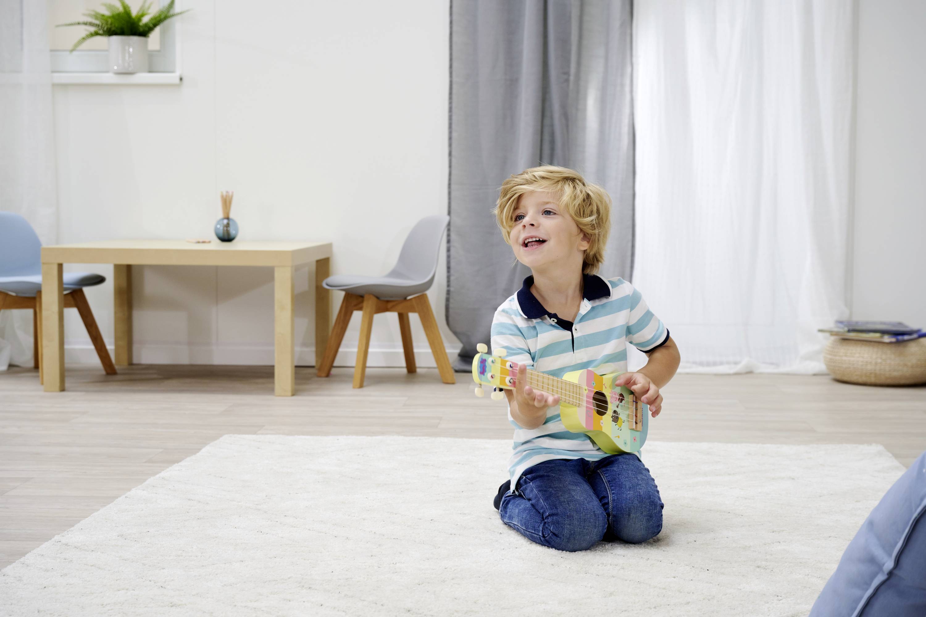 Een lachende jongen knielt op een tapijt en speelt een kleurrijke ukelele. Op de achtergrond zijn een tafel, stoelen en planten te zien.