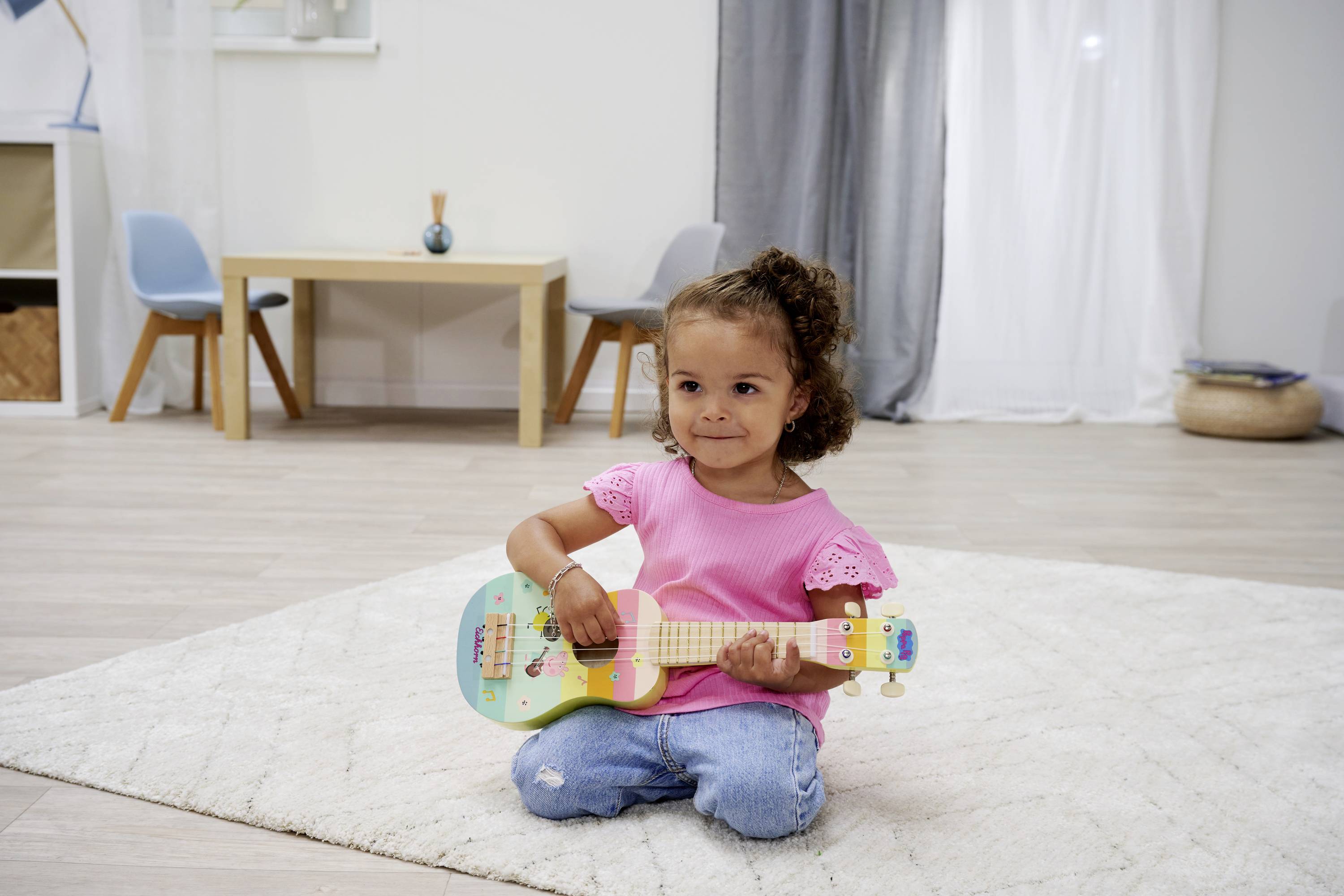 Een kind zit op een tapijt en speelt een kleurrijk speelgoedinstrument. Op de achtergrond zijn een tafel, stoelen en gordijnen te zien.