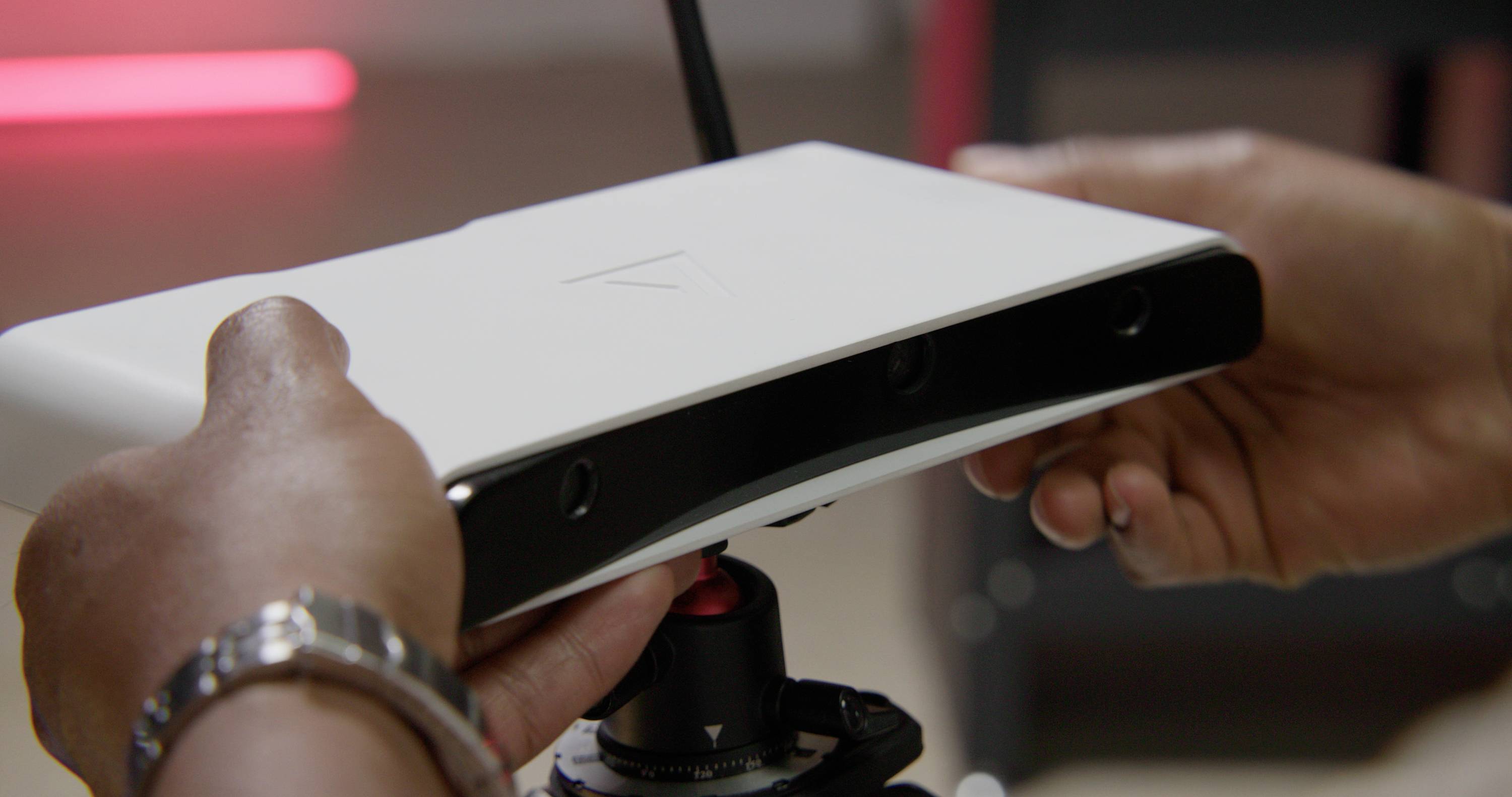 Twee handen houden een technisch apparaat met meerdere lenzen vast, mogelijk een camera of scanner, boven een statief in een binnenruimte.