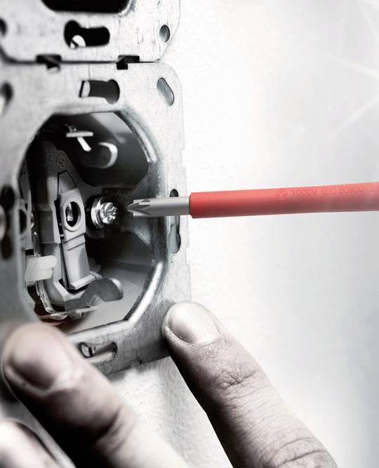 Een persoon draait met een rode schroevendraaier aan een metalen beugel, waarschijnlijk een stopcontact of schakelaar aan het installeren.