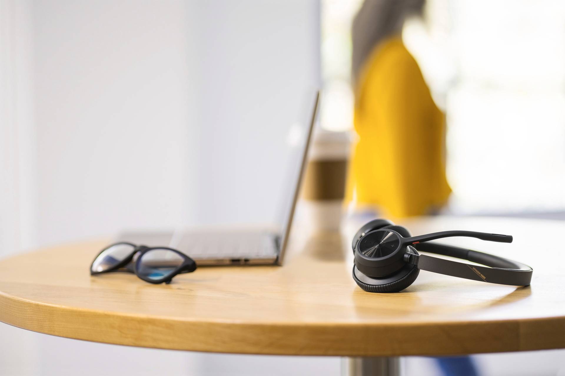 Een laptop, een bril en een headset liggen op een rond houten tafel. Op de achtergrond is een persoon wazig te zien.