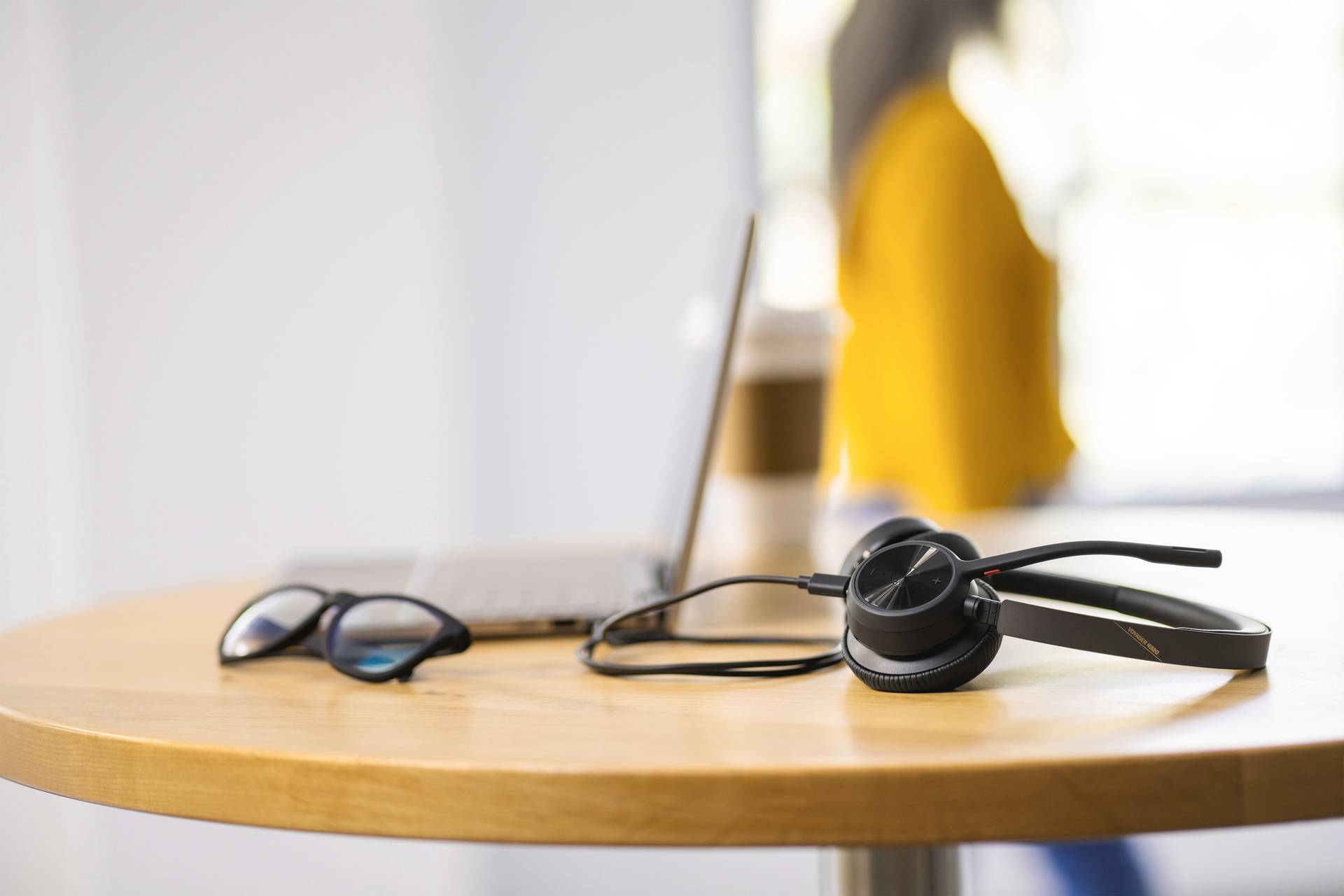 Op een rond tafeltje liggen een laptop, een bril en een headset. Op de achtergrond draagt een persoon een geel bovenkleding.