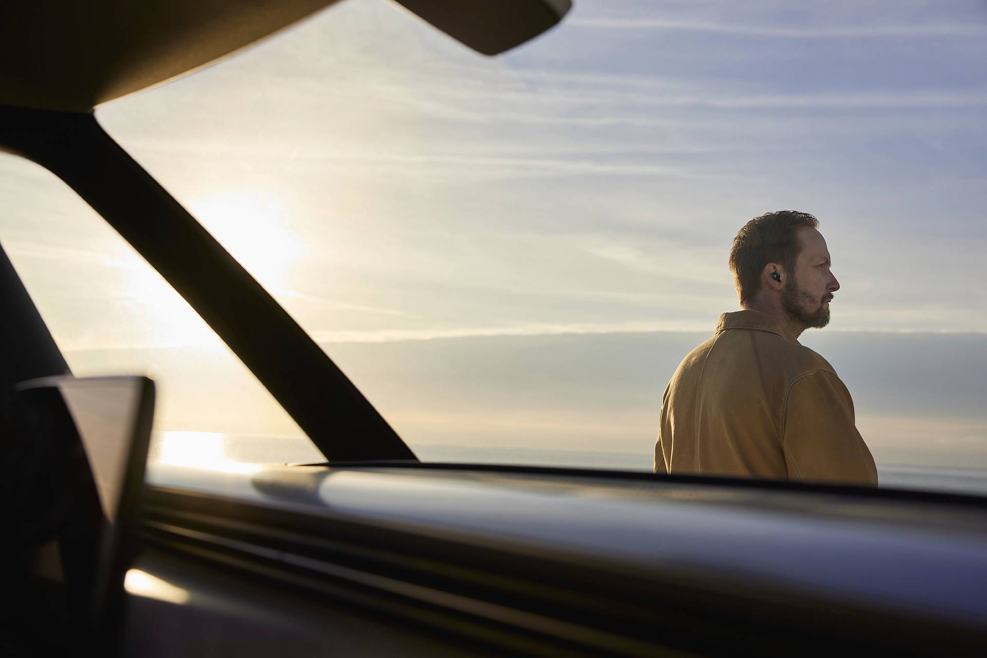 Een man staat naast een auto bij zonsondergang en kijkt in de verte terwijl hij oordopjes draagt.