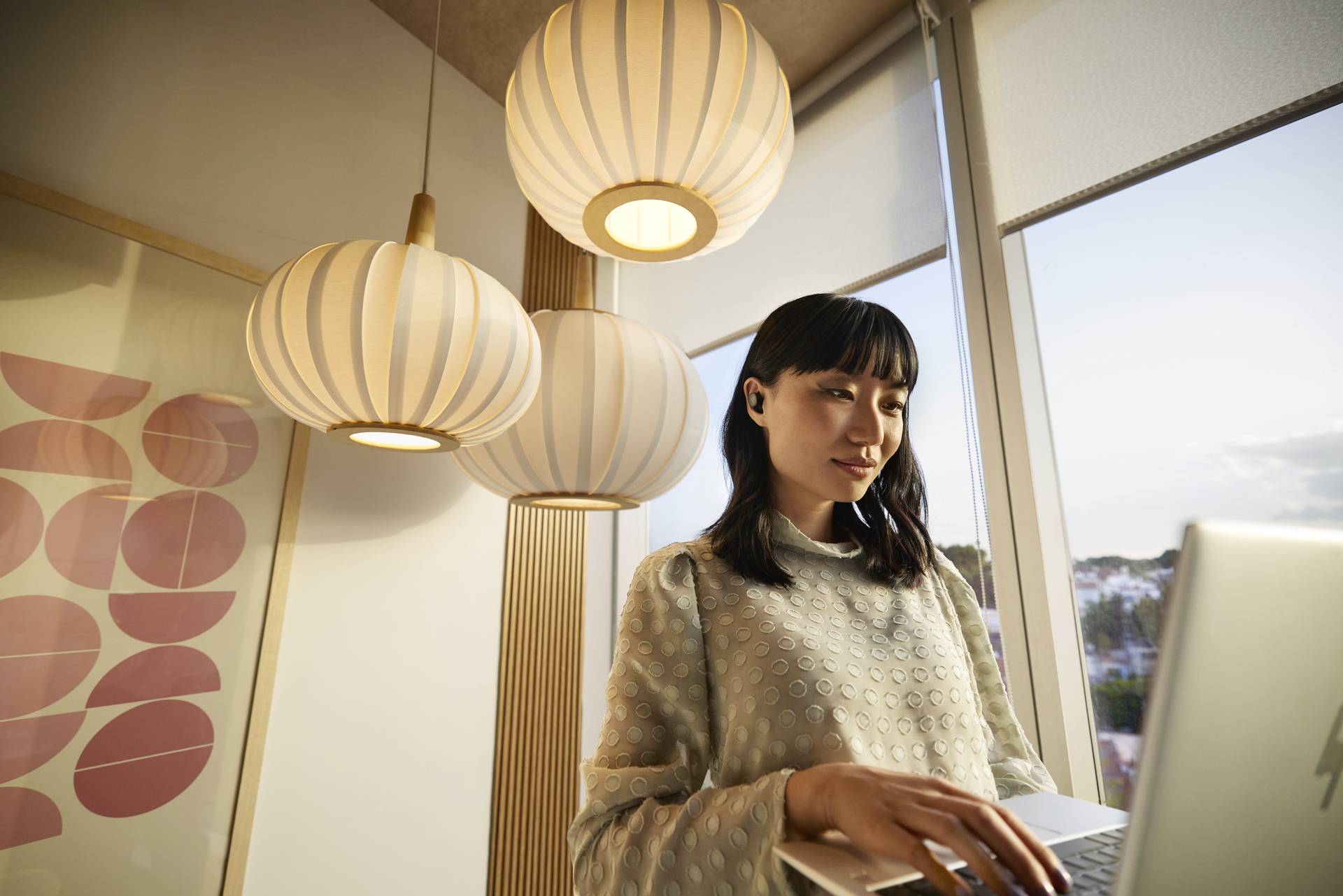 Een persoon werkt aan een laptop in een gezellig verlichte ruimte met grote, ronde hangende lampen en een raam op de achtergrond.