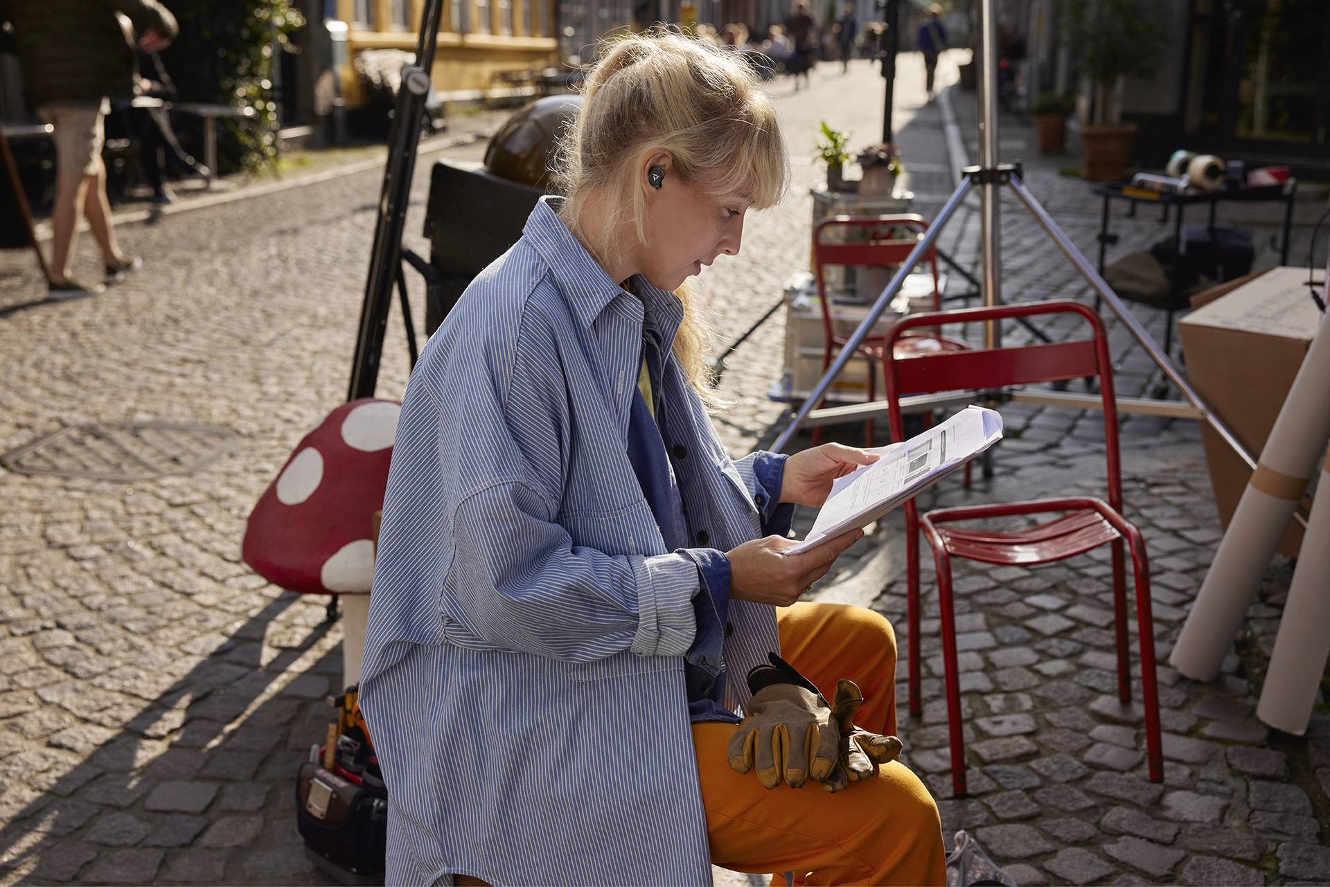 Een vrouw zit buiten op een bestrate weg en leest een document. Ze draagt koptelefoons en werkkleding. Op de achtergrond zijn straatmeubilair en bestrating zichtbaar.