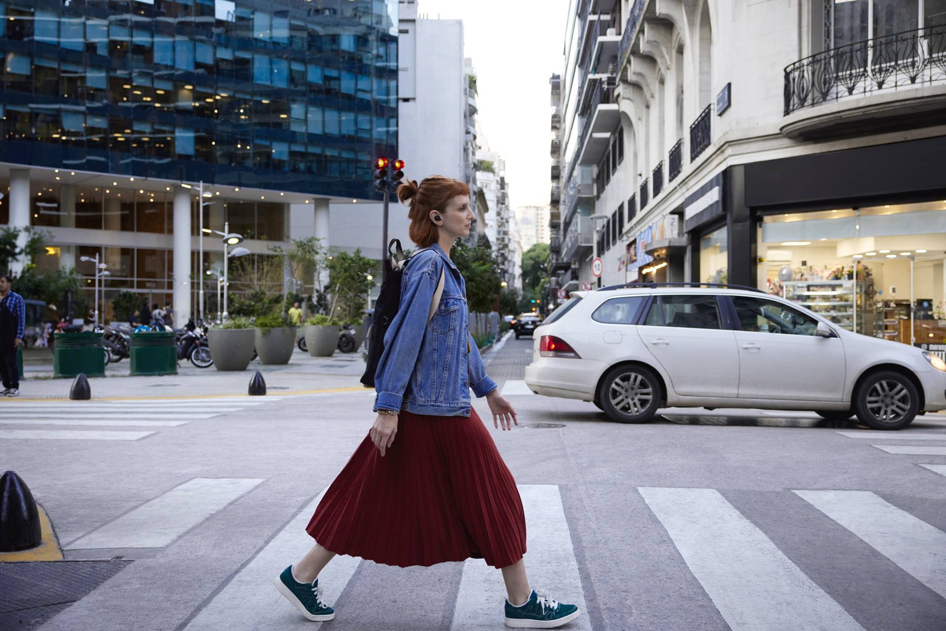 Een vrouw met een rode rok en een spijkerjasje steekt overdag een stadsstraat over in de zebrapadzone. Gebouwen en auto's op de achtergrond.