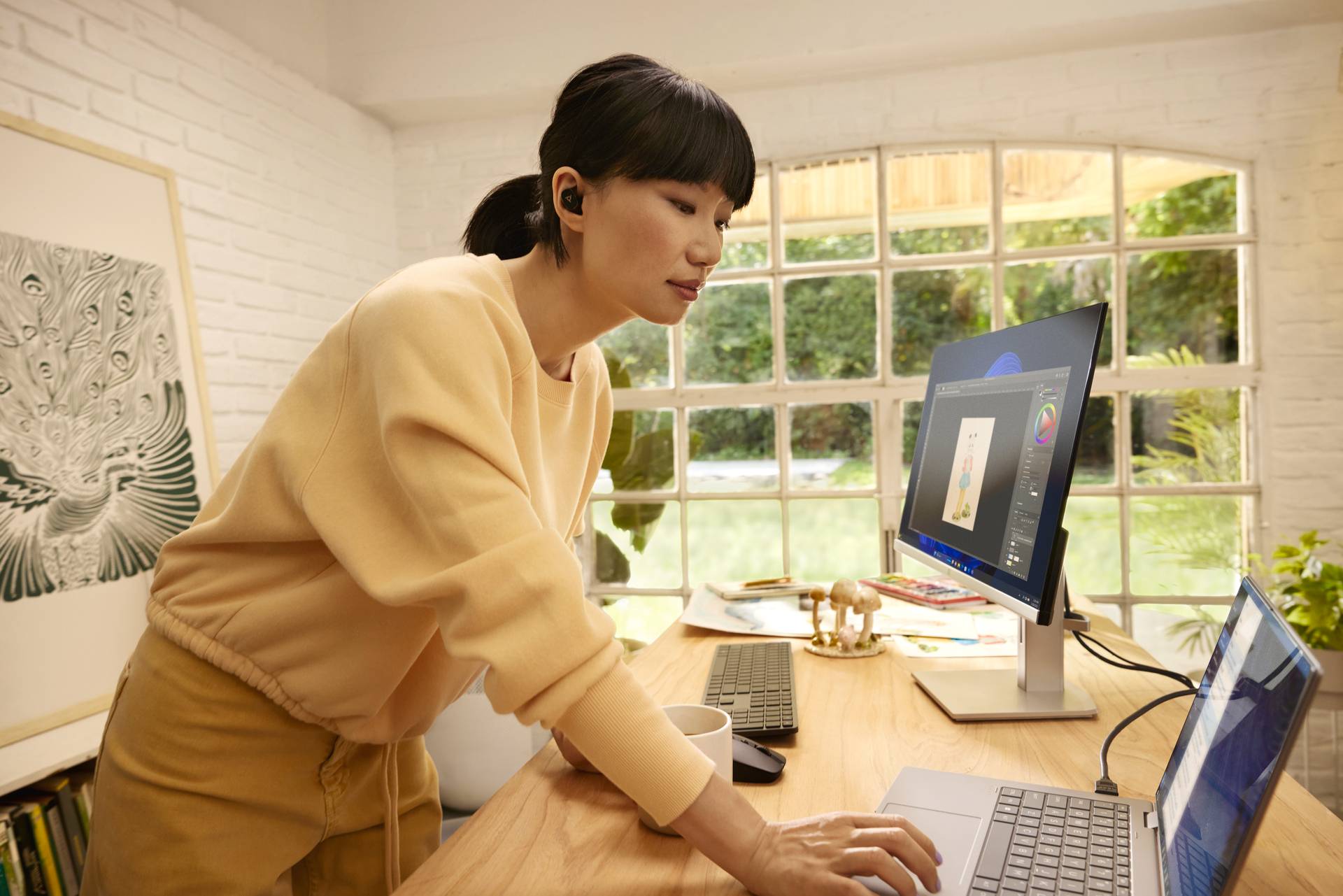 Een vrouw werkt aan een bureau met twee schermen in een zonnige ruimte. Ze draagt lichte kleding en heeft donker haar.