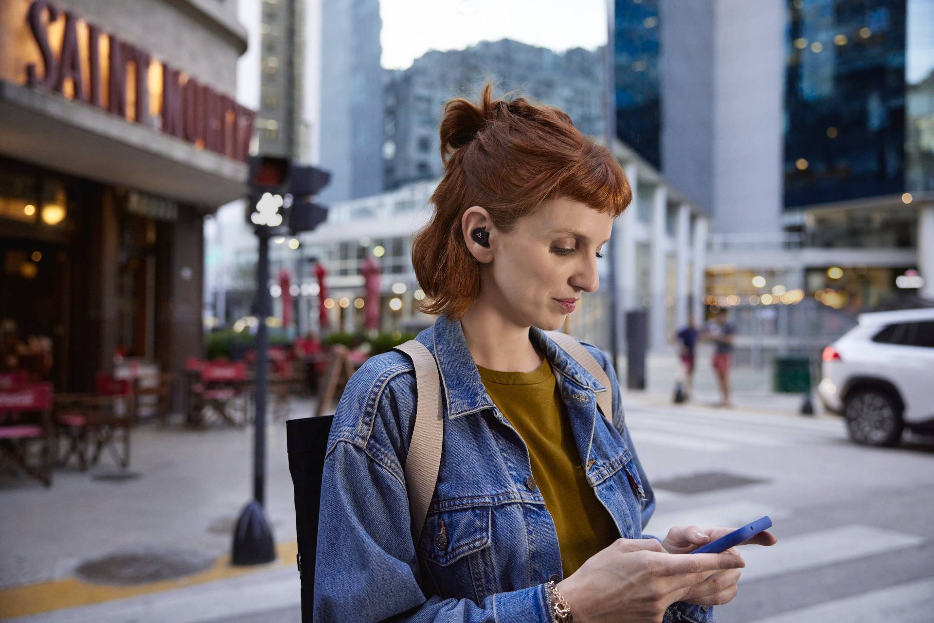 Een vrouw met kort rood haar en oordopjes staat aan een stadsstraat en kijkt naar haar smartphone. Op de achtergrond zijn gebouwen zichtbaar.