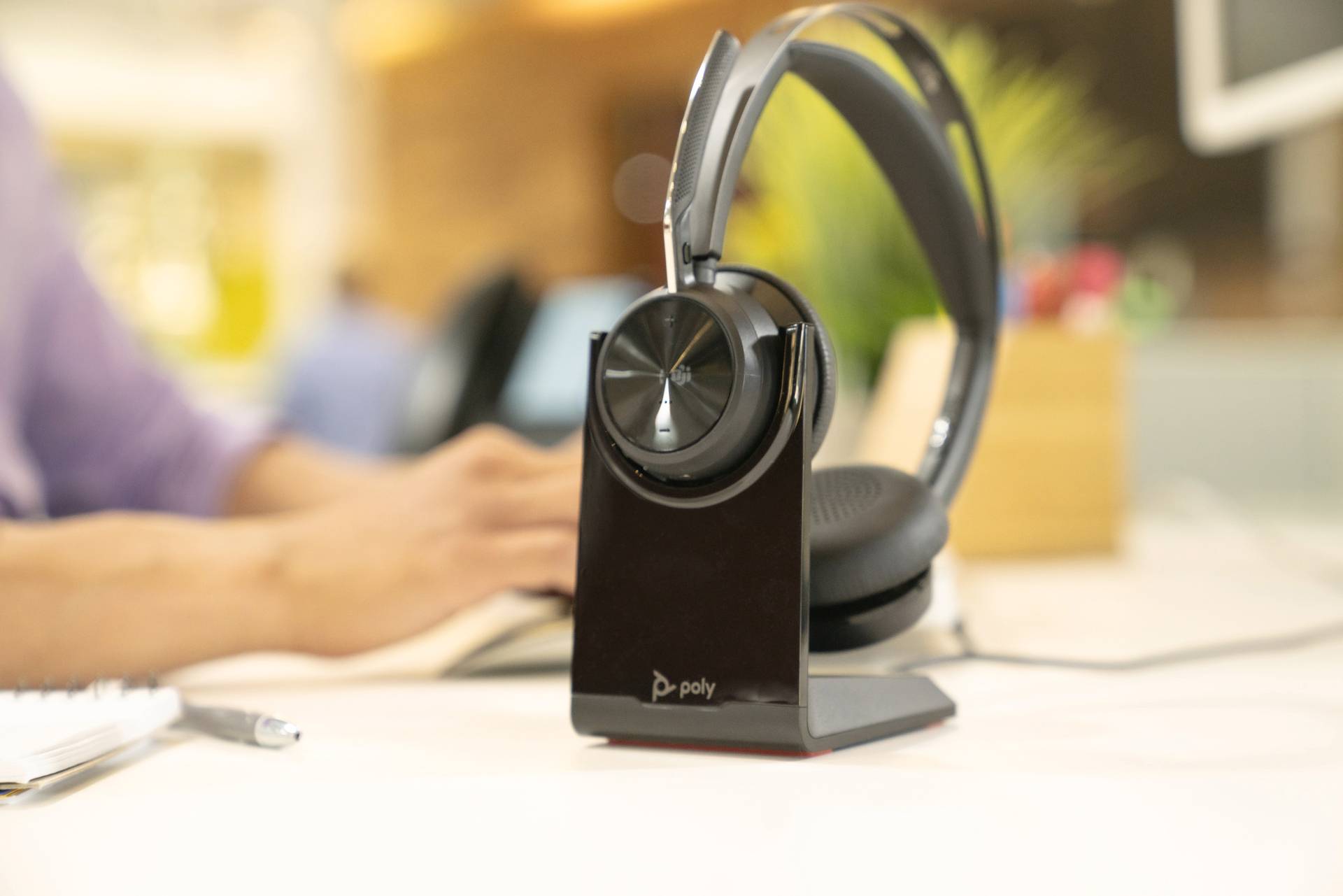 Een headset op een tafel met laadstation. Op de achtergrond typt iemand op een toetsenbord. Kantooromgeving met planten.
