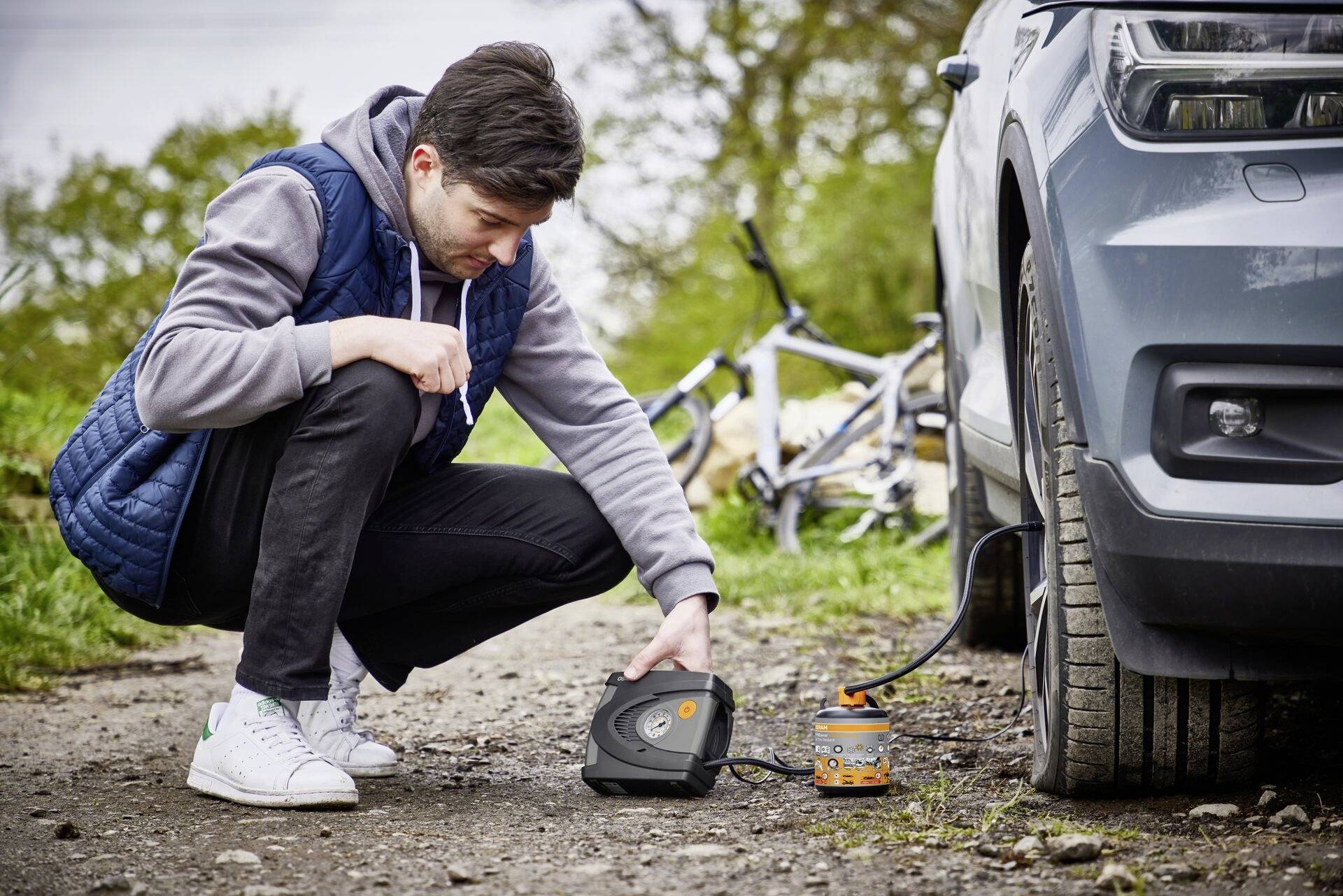 Een man knielt op een onverharde weg en gebruikt een draagbare compressor om de band van een auto op te pompen. Op de achtergrond staat een fiets.