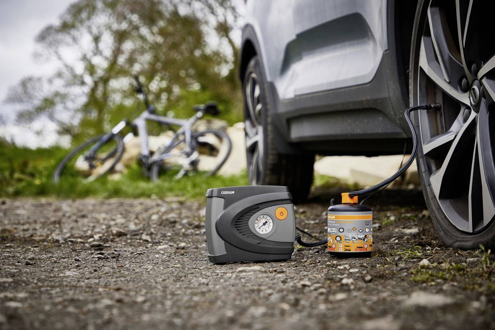 Een autoband wordt opgepompt met een draagbare compressor, op de achtergrond staat een fiets op een veldweg.