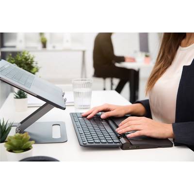 Een persoon werkt aan een bureau met een laptopstandaard en toetsenbord; op de achtergrond zit nog een persoon aan een vergelijkbare werkplek.