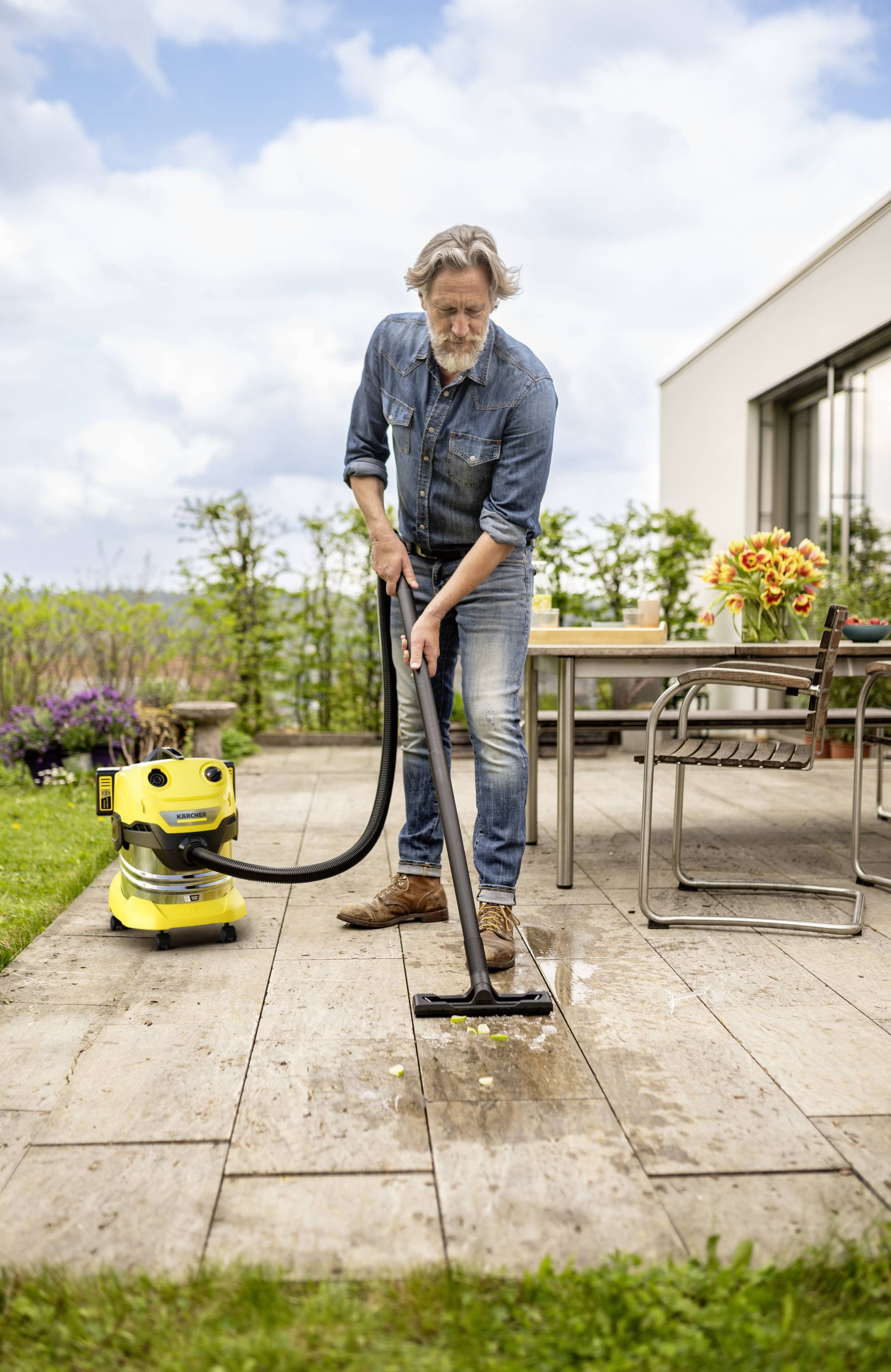 Een man reinigt een houten terras met een gele industriestofzuiger. Op de achtergrond zijn planten en een tafel met bloemen.