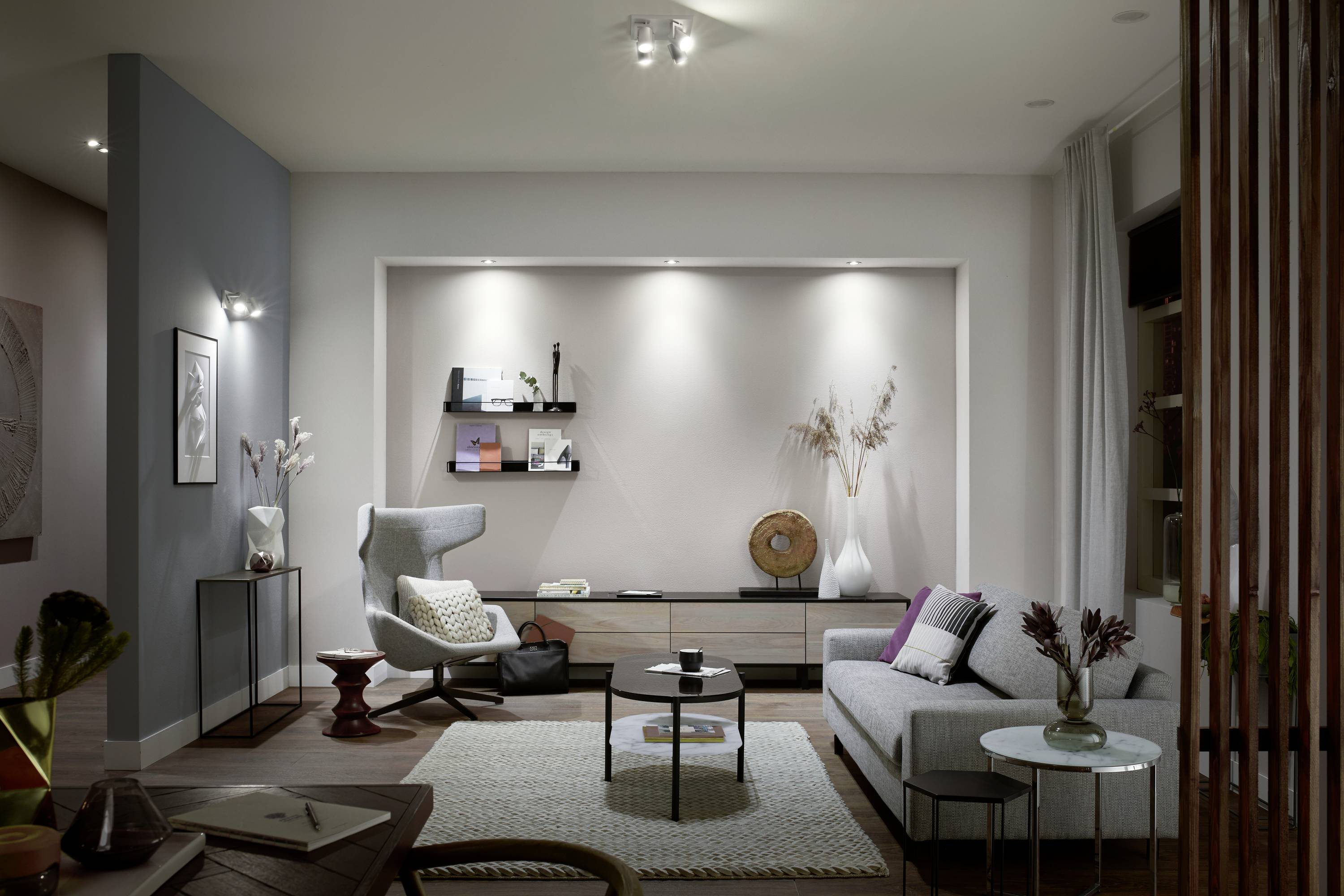 Moderne woonruimte met een grijs bankstel, bijzettafel, fauteuil en subtiele decoraties. Kunstwerken aan de muur, neutrale kleuren.