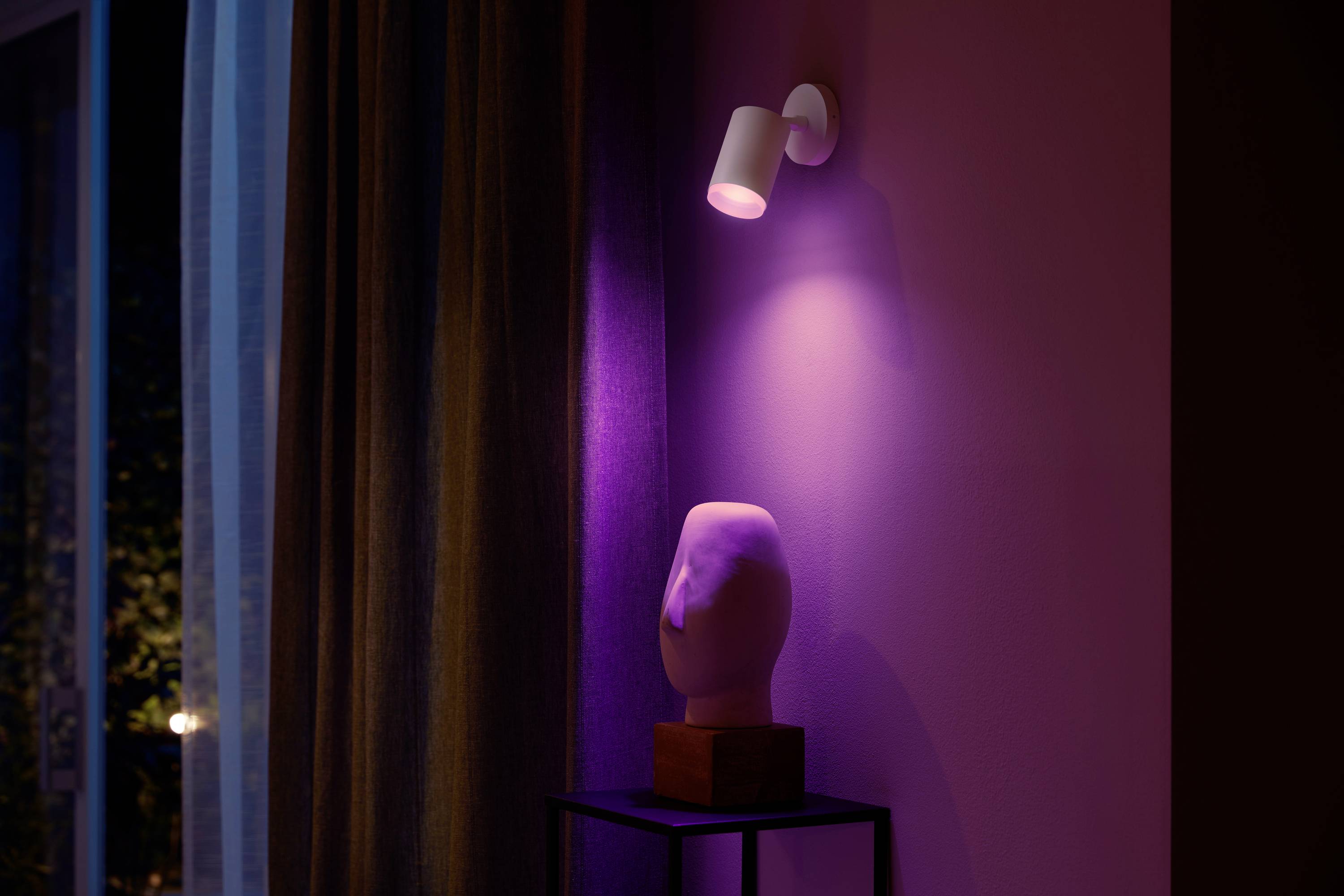 Een witte schijnwerper verlicht een abstracte sculptuur op een tafel in een ruimte met paars licht en een gordijn op de achtergrond.