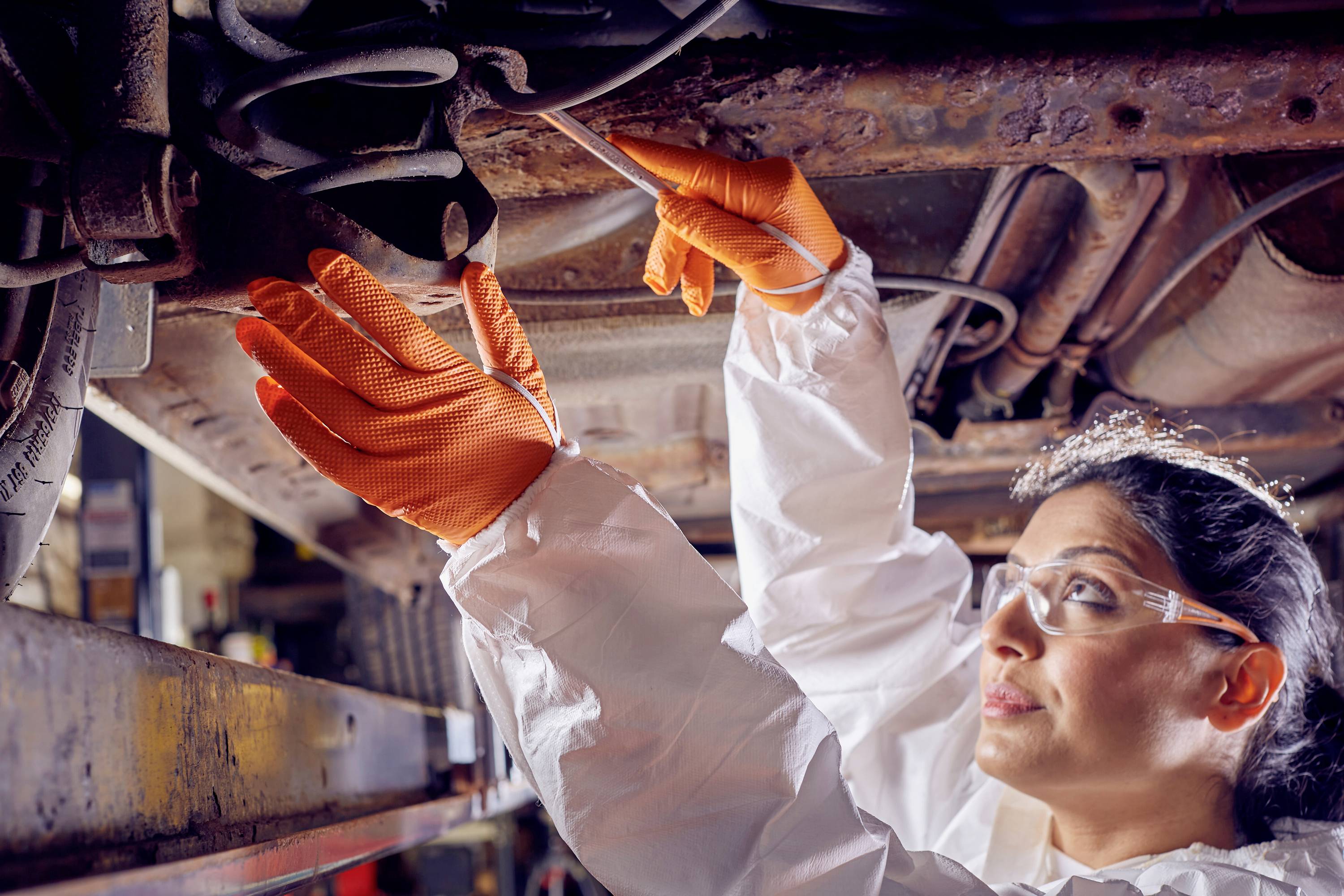 Een monteur in beschermende kleding en handschoenen inspecteert de onderkant van een voertuig in een autogarage.