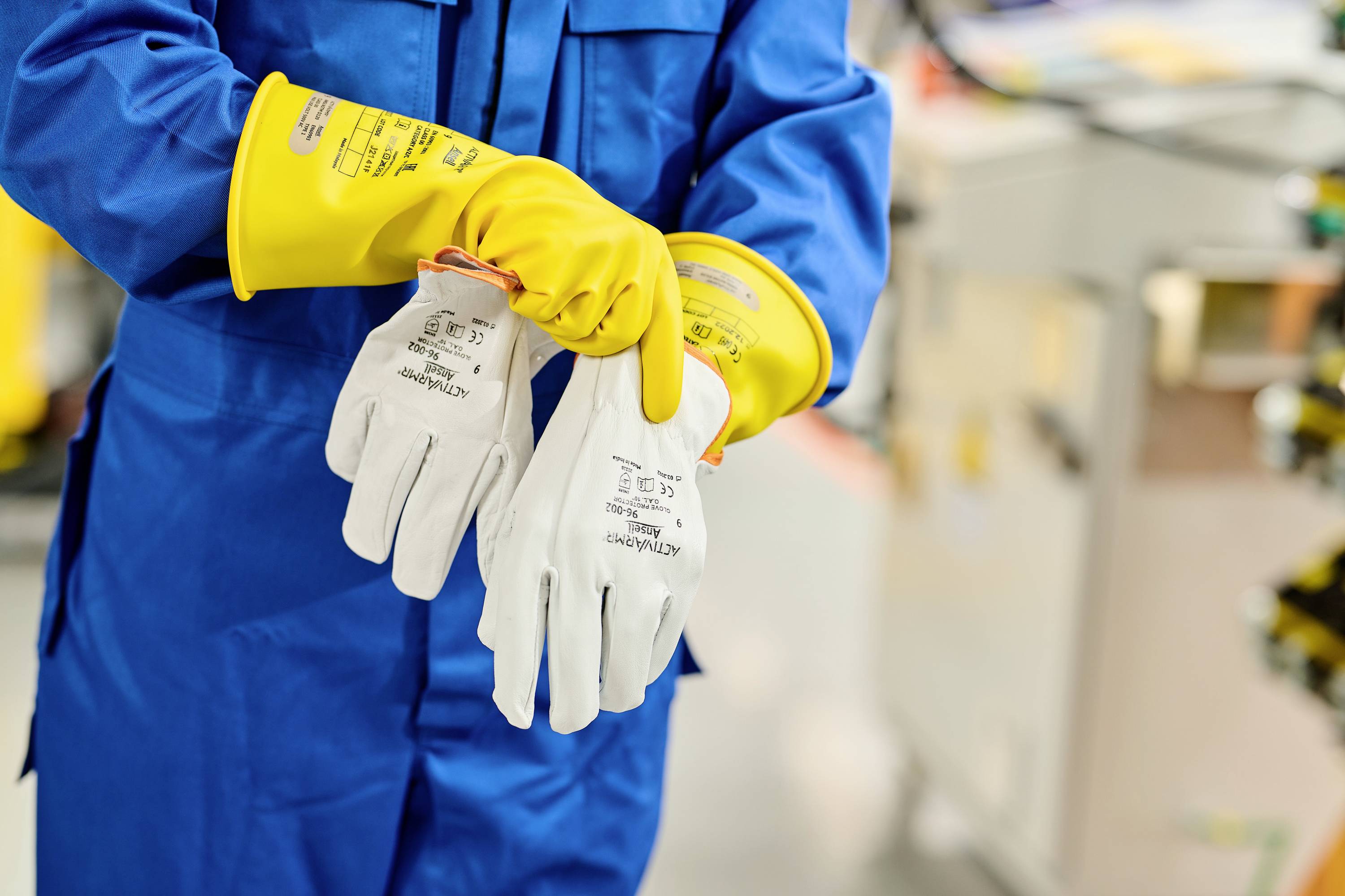 Een persoon in blauwe werkkleding trekt gele beschermende handschoenen over witte isolatiehandschoenen aan om veiligheid te waarborgen.