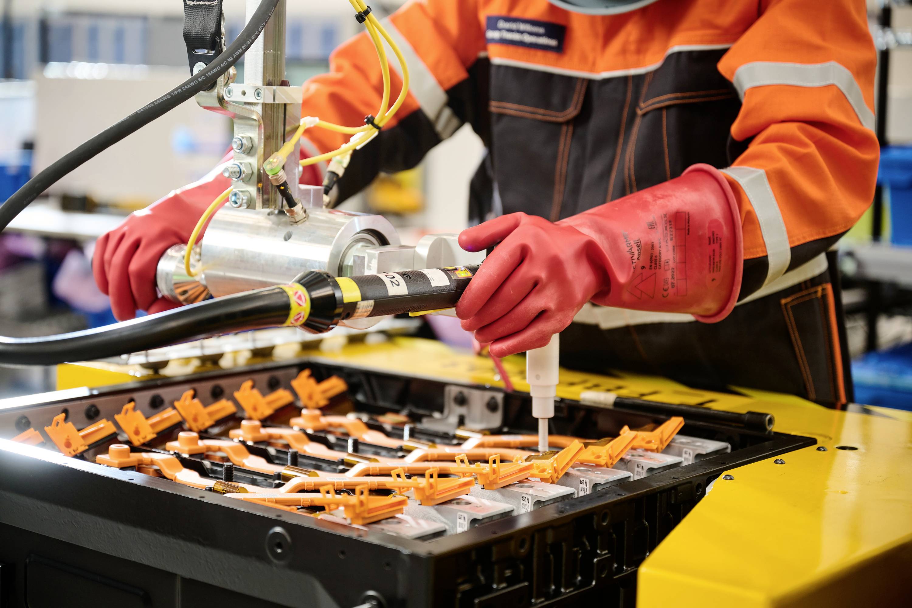 Een persoon in beschermende kleding en rode handschoenen werkt aan een batterij. Ze gebruikt een gereedschap om verbindingen te testen.