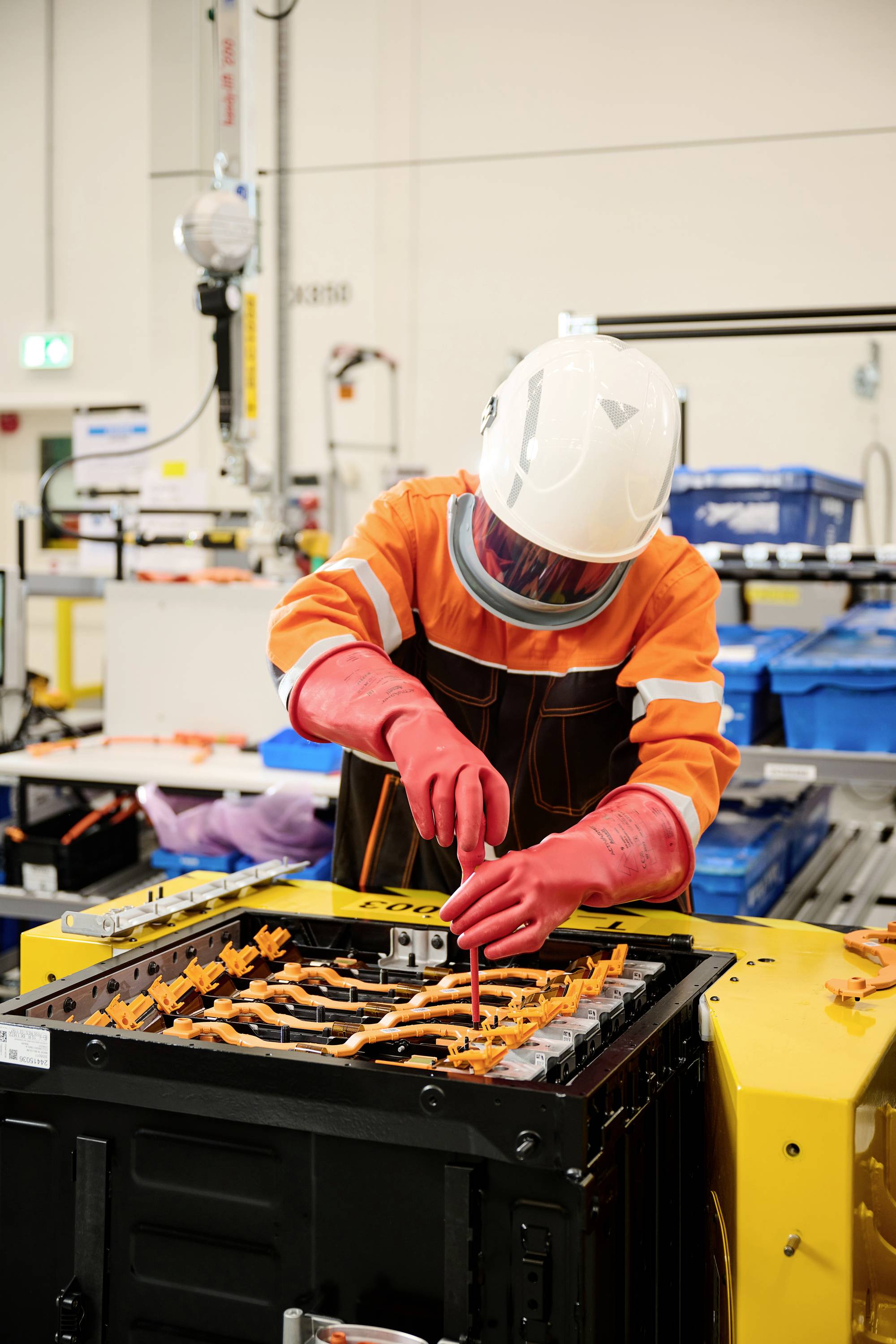 Arbeider in beschermende kleding monteert een batterij in een industriële omgeving. Verschillende gereedschappen en onderdelen zijn op de achtergrond zichtbaar.