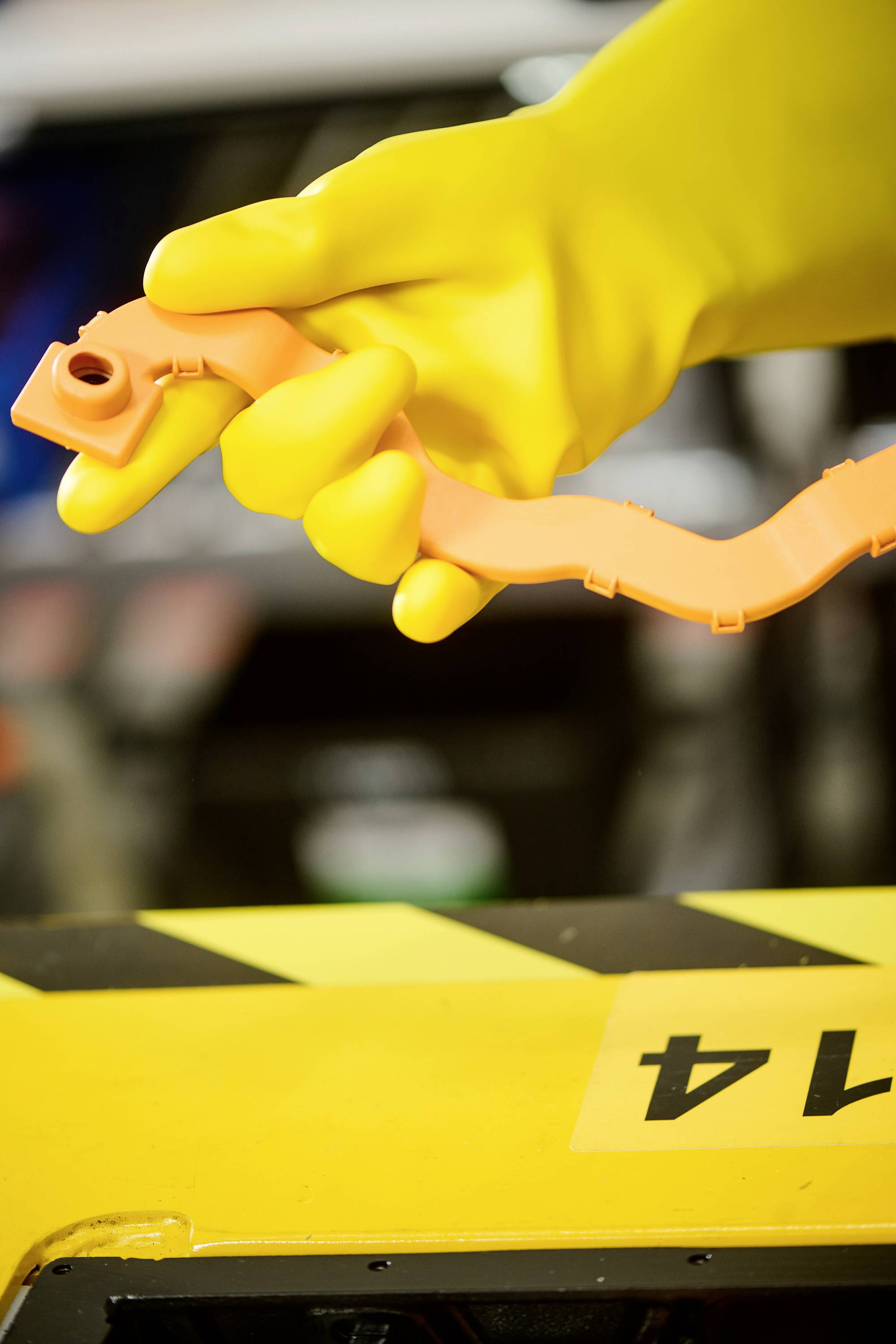 Een hand in een gele rubberen handschoen houdt een oranje gereedschap vast boven een geel oppervlak met zwarte strepen. Op de achtergrond een onscherp voertuig.