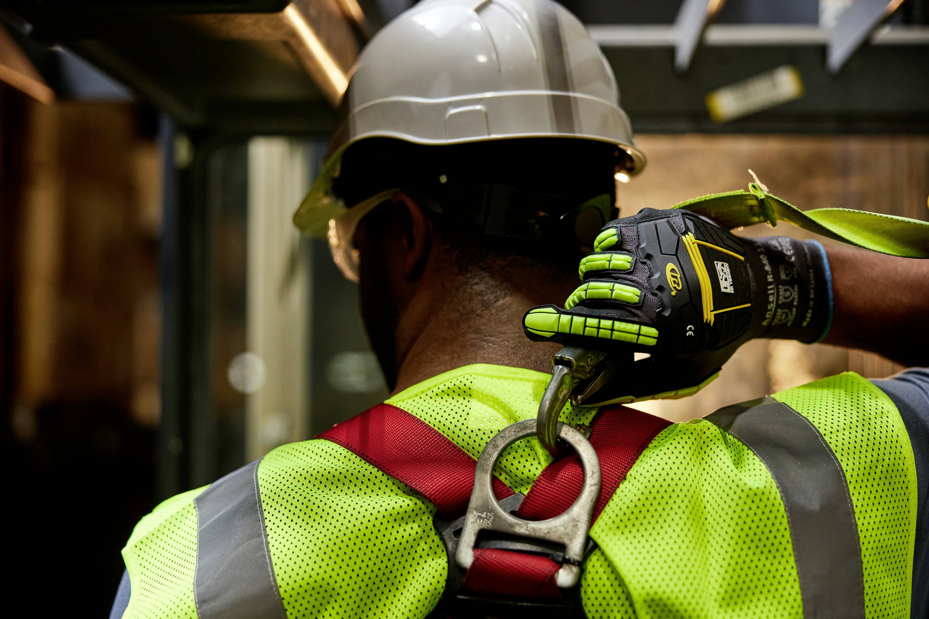Een persoon draagt beschermende kleding en een helm terwijl hij/zij aan het werk is op de bouwplaats. Hij/zij bevestigt een veiligheidsharnas aan een ankerpunt.