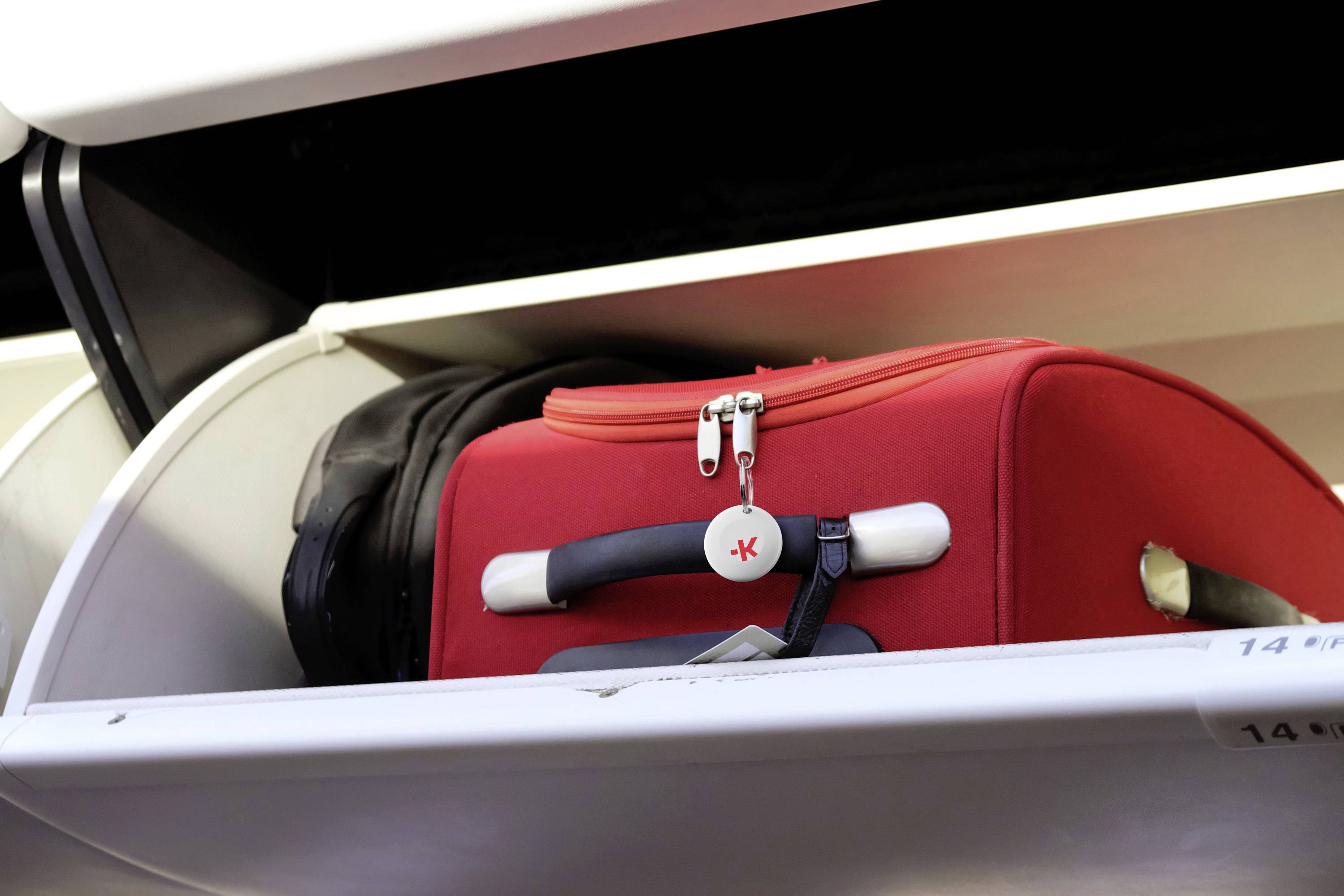 Rode koffer met etiket opgeborgen in een bagagevak voor handbagage in een vliegtuig. Ernaast een opgevouwen jas.