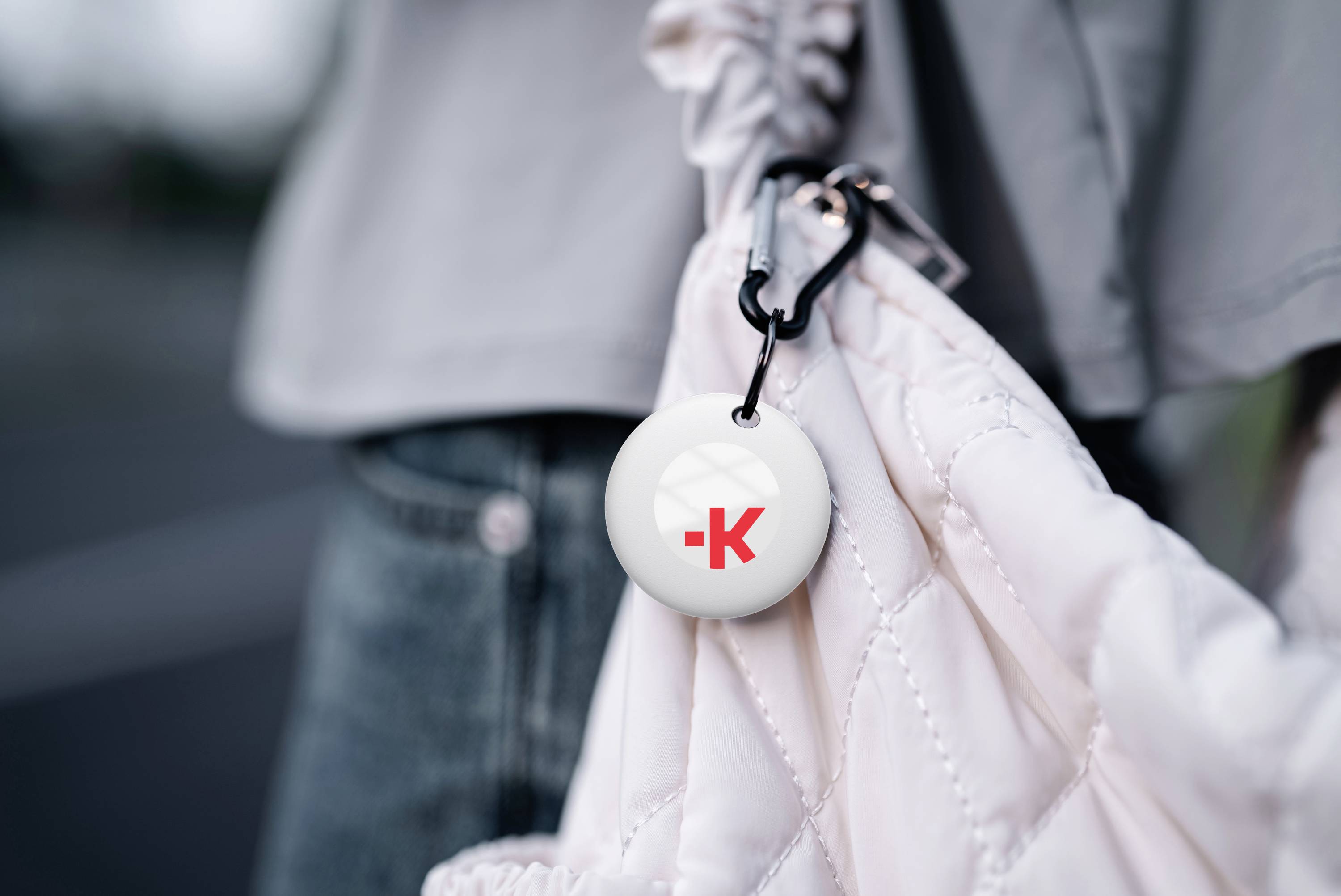 Een sleutelhanger met een rood 'K'-logo hangt aan een witte gewatteerde tas. De persoon draagt een grijze jas.