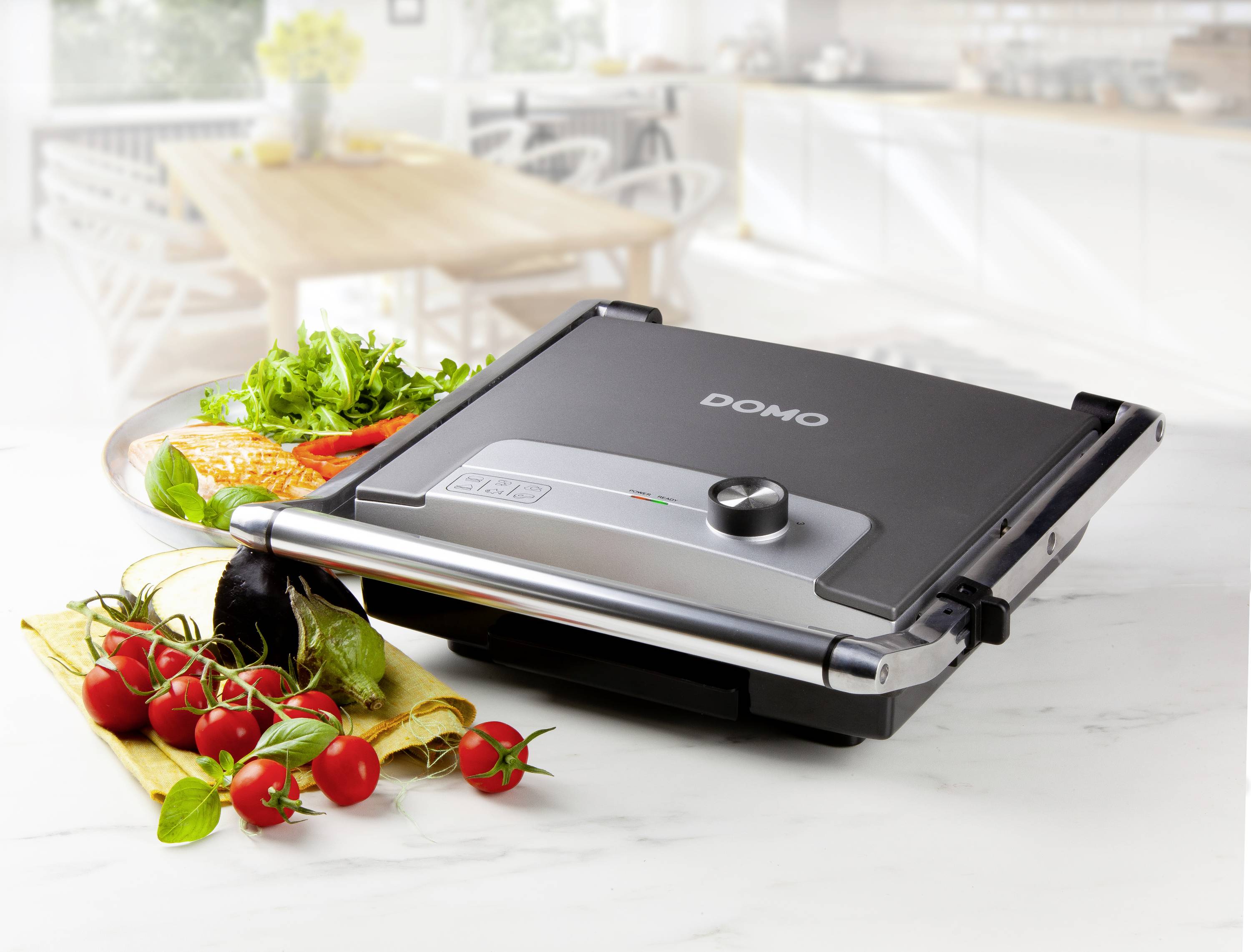 Een contactgrill op een keukenaanrecht, omringd door verse ingrediënten zoals tomaten, paprika's en kruiden. Op de achtergrond is een keuken zichtbaar.