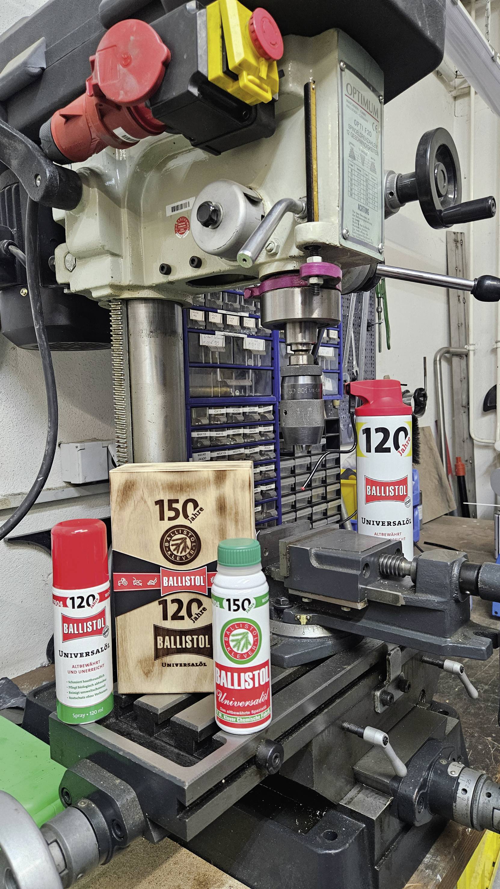 Boormachine in een werkplaats met meerdere Ballistol-producten op de voorgrond, inclusief spuitbussen en houten verpakking.