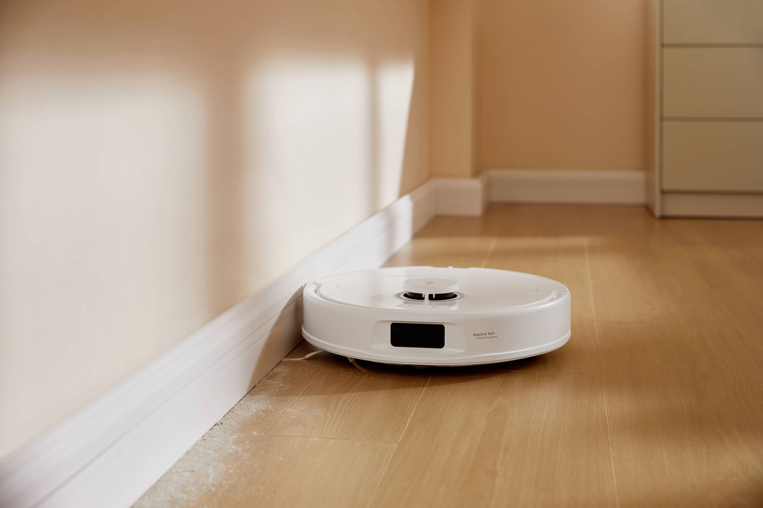 Een witte robotstofzuiger reinigt de houten vloer langs een muur in een lichte ruimte.