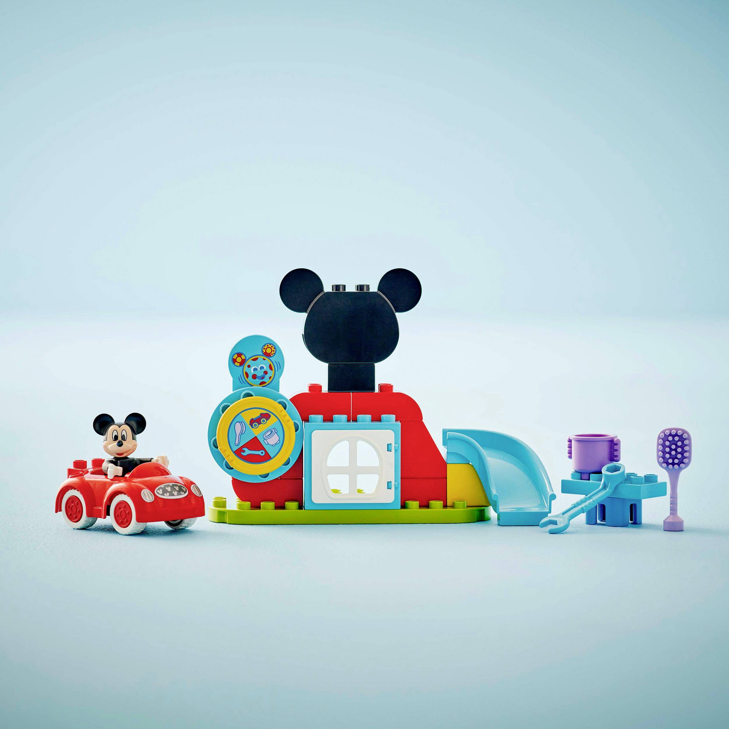 Een speelgoedset met Mickey Mouse in een rode auto, een kleurrijk blokkenhuis met schep, emmer en tandenborstel voor een blauwe achtergrond.