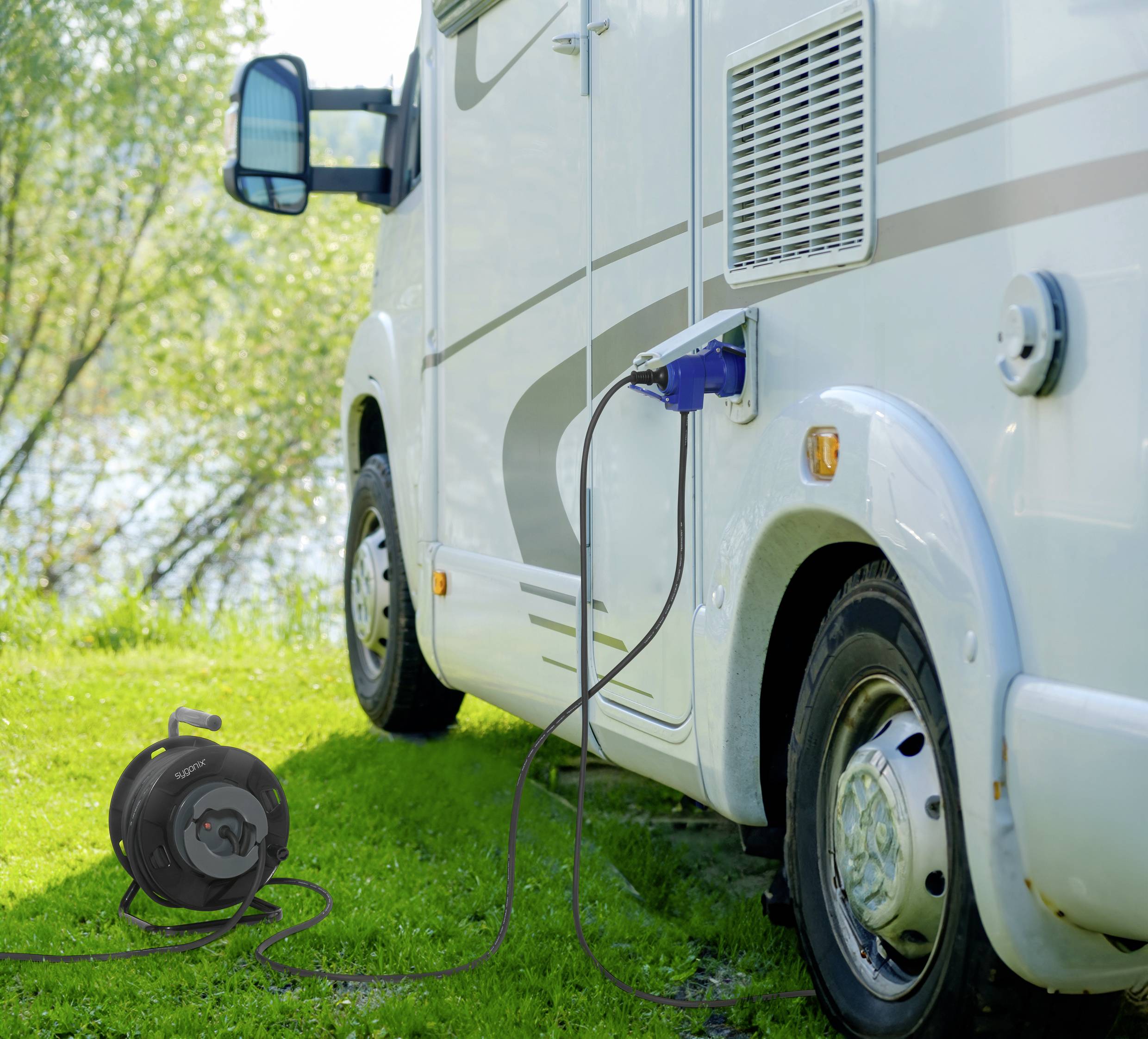 Een camper is geparkeerd op een groene weide en is via een kabel aangesloten op een stroombron. Op de achtergrond zijn bomen en een rivier te zien.