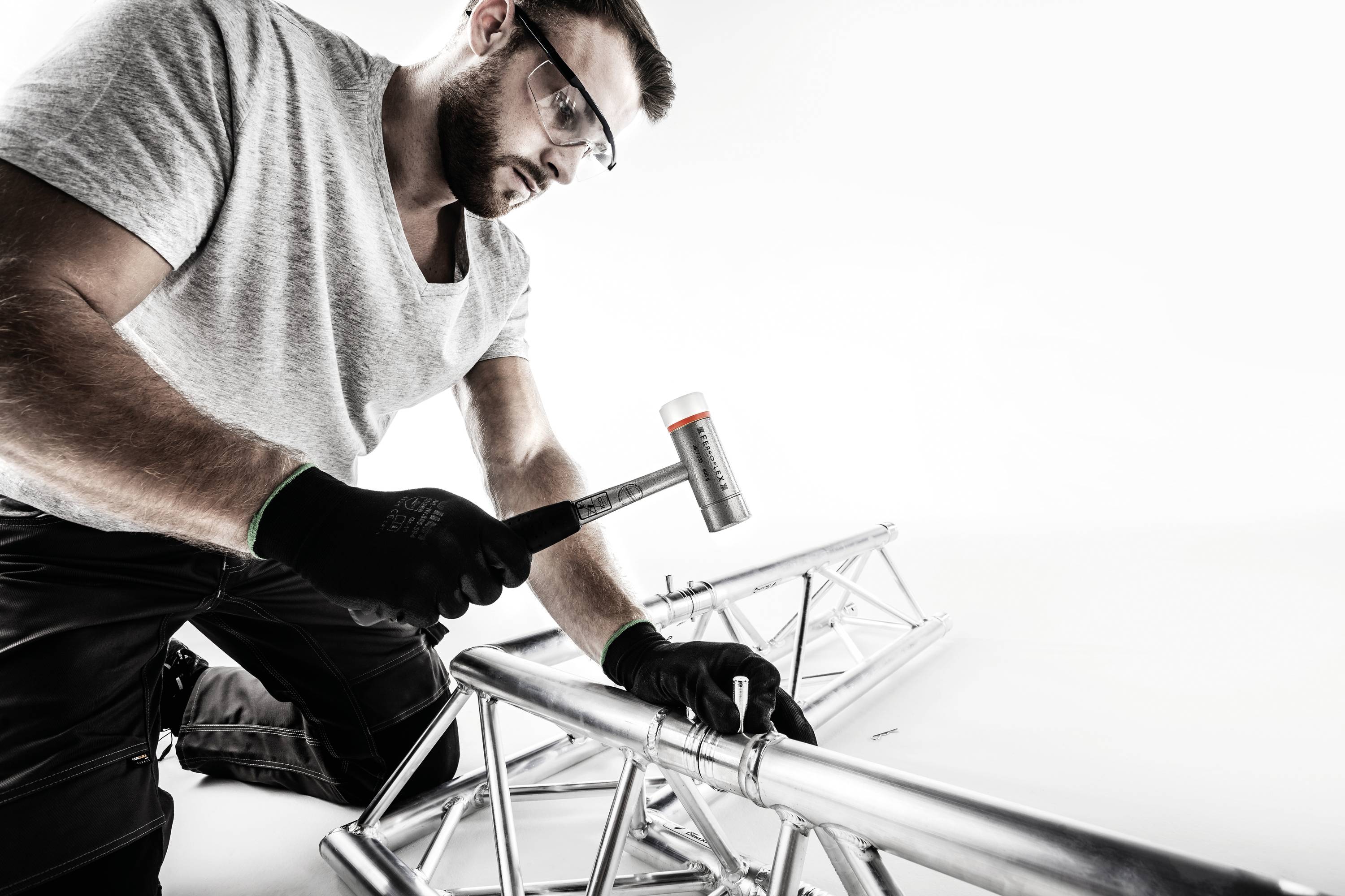 Een man werkt met een hamer aan een zilveren metalen constructie. Hij draagt een veiligheidsbril en handschoenen.