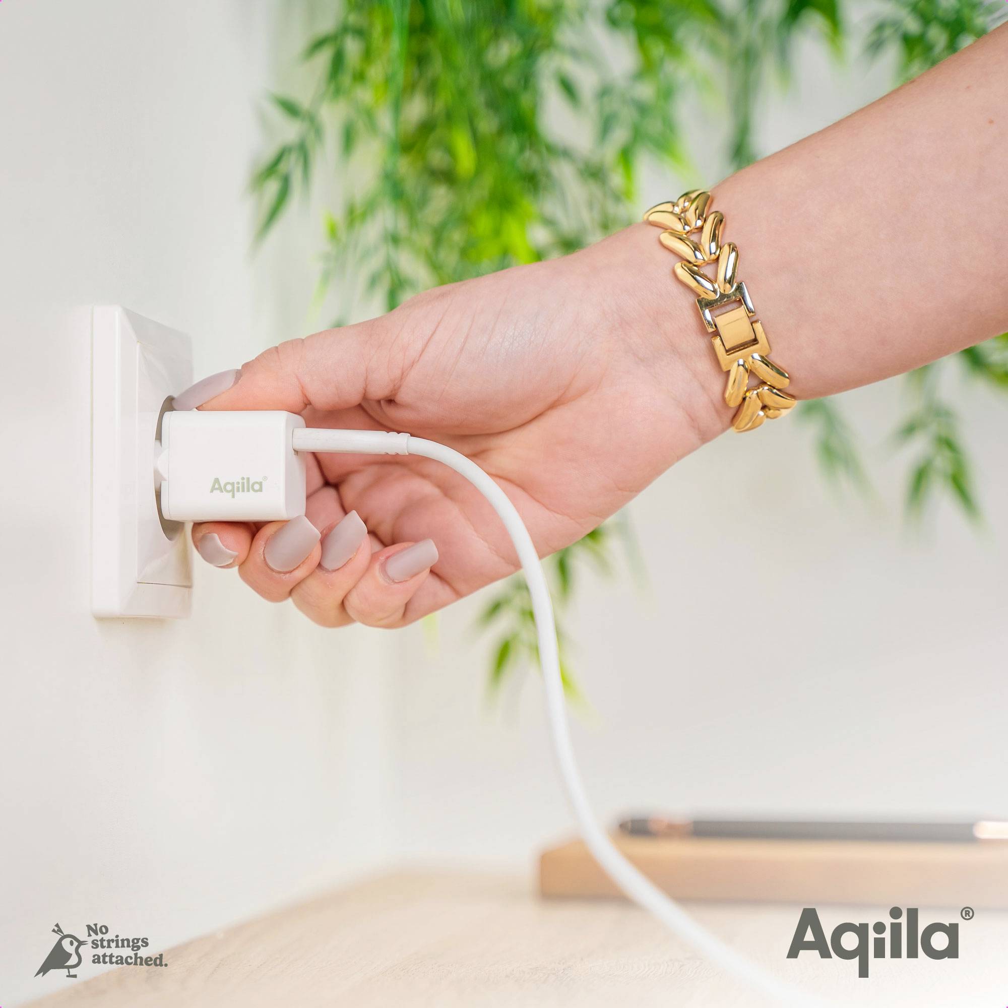 Een hand steekt een wit oplaadapparaat van 'Aqila' in een stopcontact. Op de achtergrond zijn planten zichtbaar. Op de afbeelding staat: 'No Strings Attached. Aqila'.