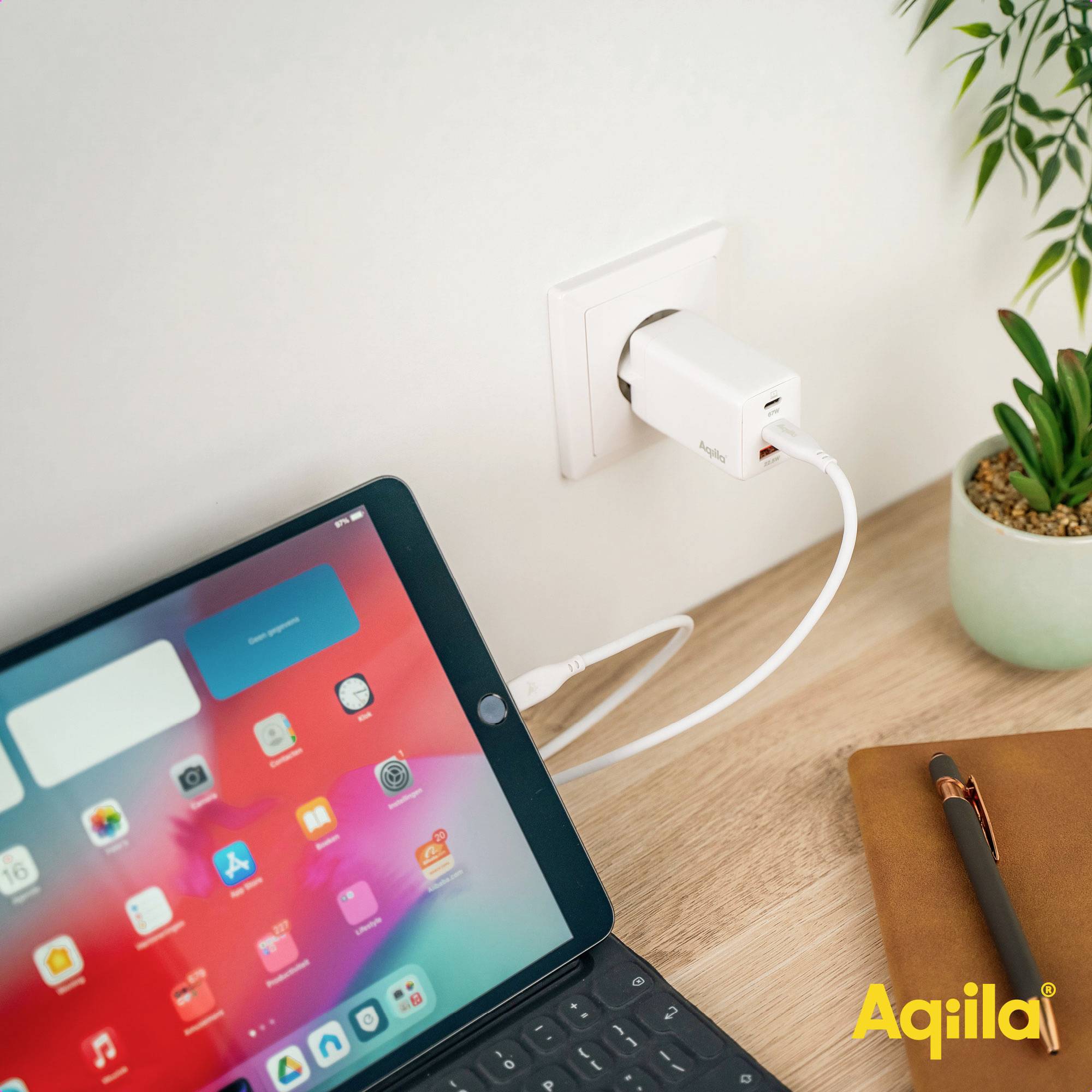 Een tablet is aangesloten op een stopcontact en wordt opgeladen. Ernaast ligt een notitieboek. Op de achtergrond staat een plant.