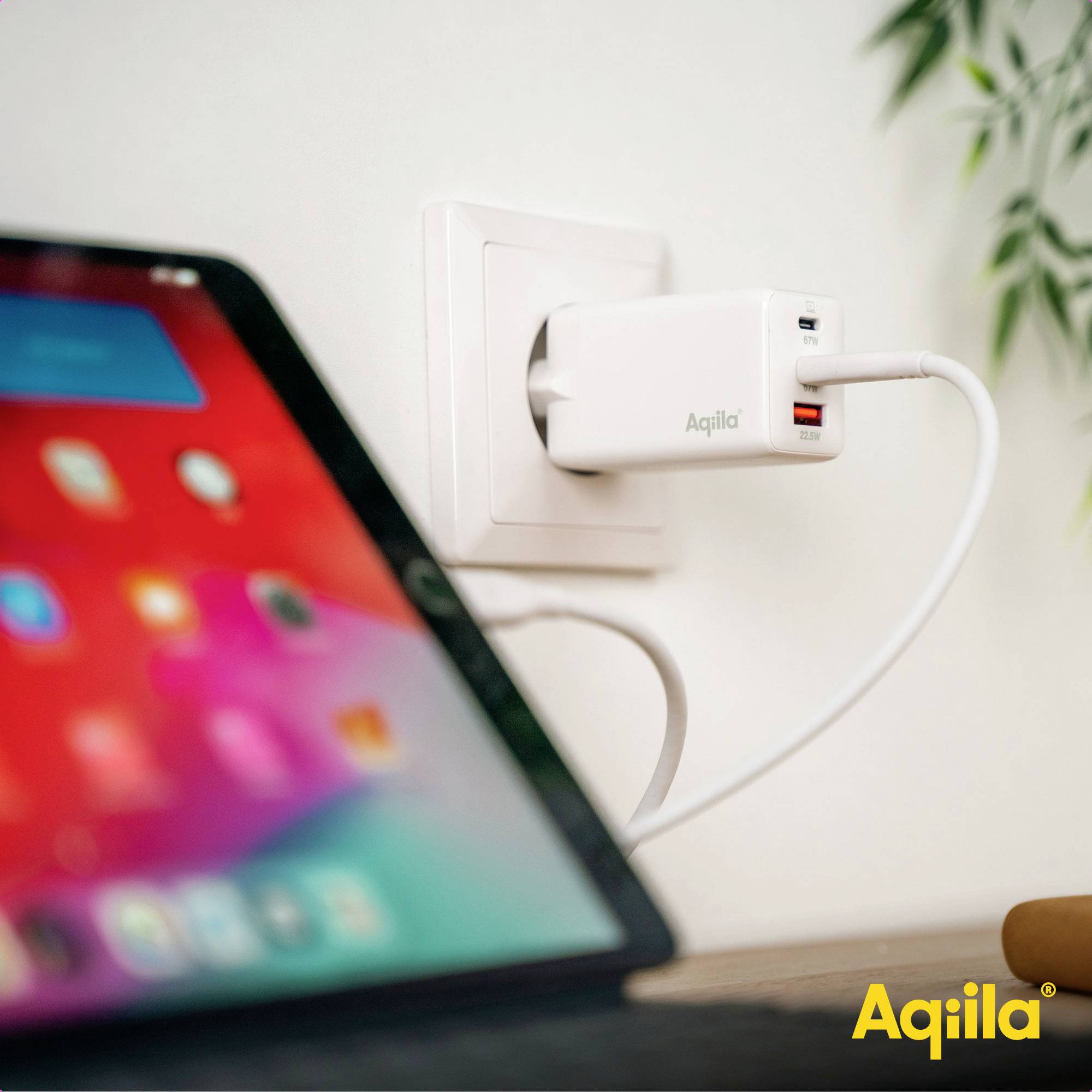Een mobiele telefoon wordt opgeladen met behulp van een Aquila-oplader die in een stopcontact is gestoken. Een tablet is zichtbaar in de buurt.