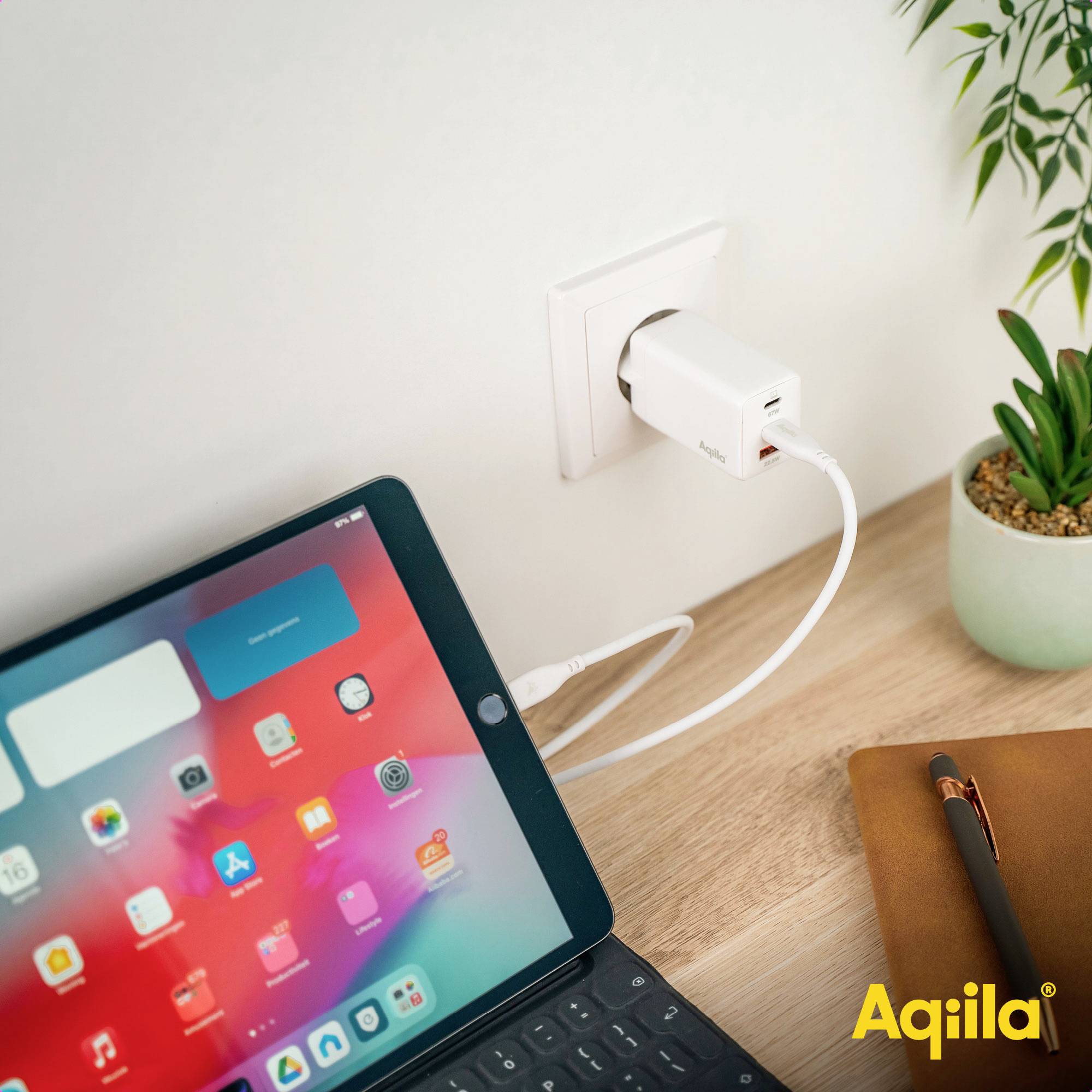 Een tablet wordt opgeladen aan een stopcontact op een houten tafel, naast een notitieboek en een plant.