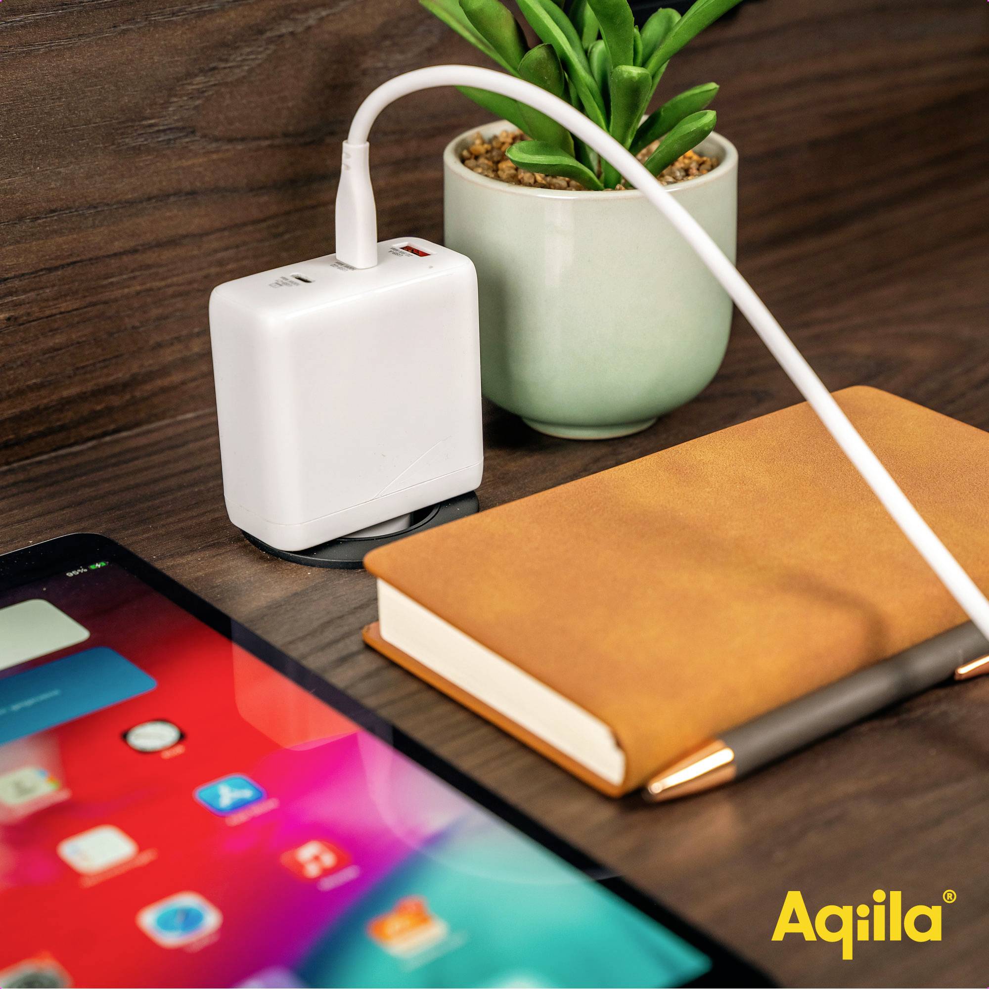 Een oplader en een notitieboek op een tafel naast een tablet. Een groene plant staat ernaast. Branding-logo 'Aqiila' in de hoek.