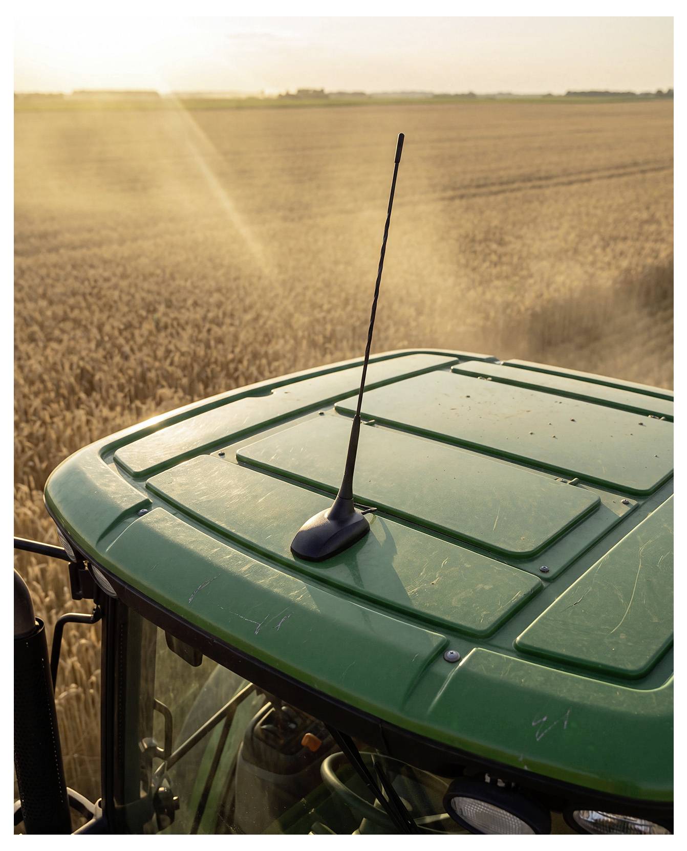 Groene tractor met cabinedak en antenne die door een gouden graanveld bij zonsondergang rijdt, van bovenaf gezien.