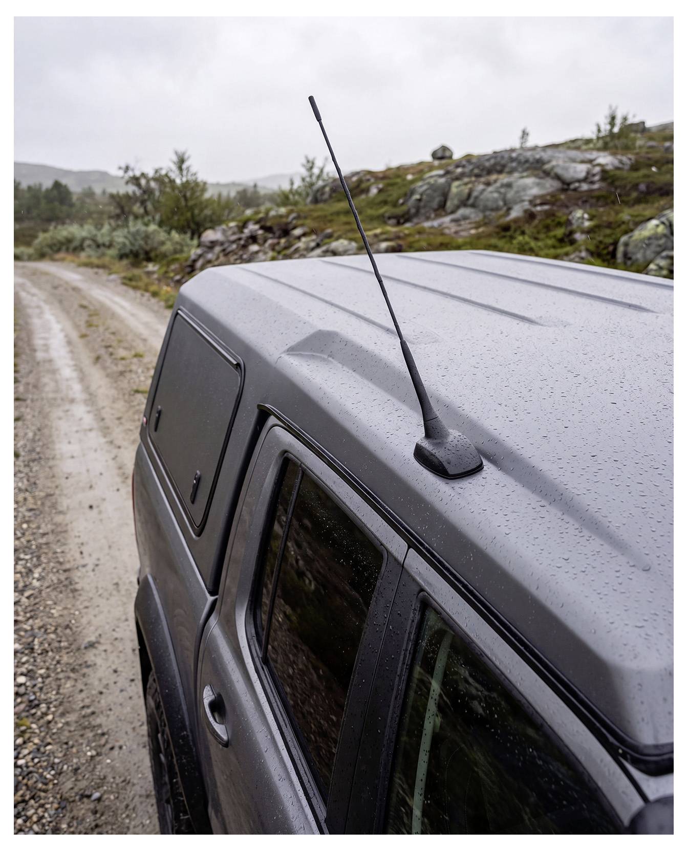 Bovenkant van een grijs terreinvoertuig op een natte grindroadpad, met een antenne en een bewolkt landschap op de achtergrond.