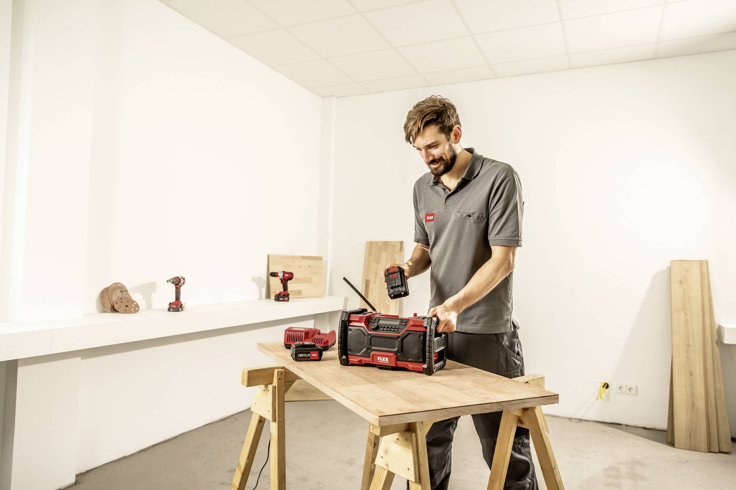 Een man staat aan een tafel en houdt een batterij vast, klaar om deze in een rood bouwradio te plaatsen. Op de achtergrond zijn gereedschappen zichtbaar.