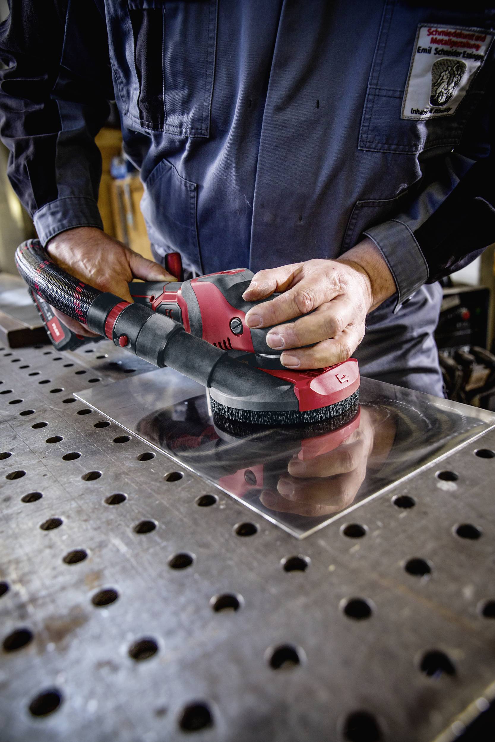 Persoon slijpt metalen plaat met een rode slijpmachine. Draagt blauwe werkkleding en staat aan een met gereedschap bezaaide werktafel.