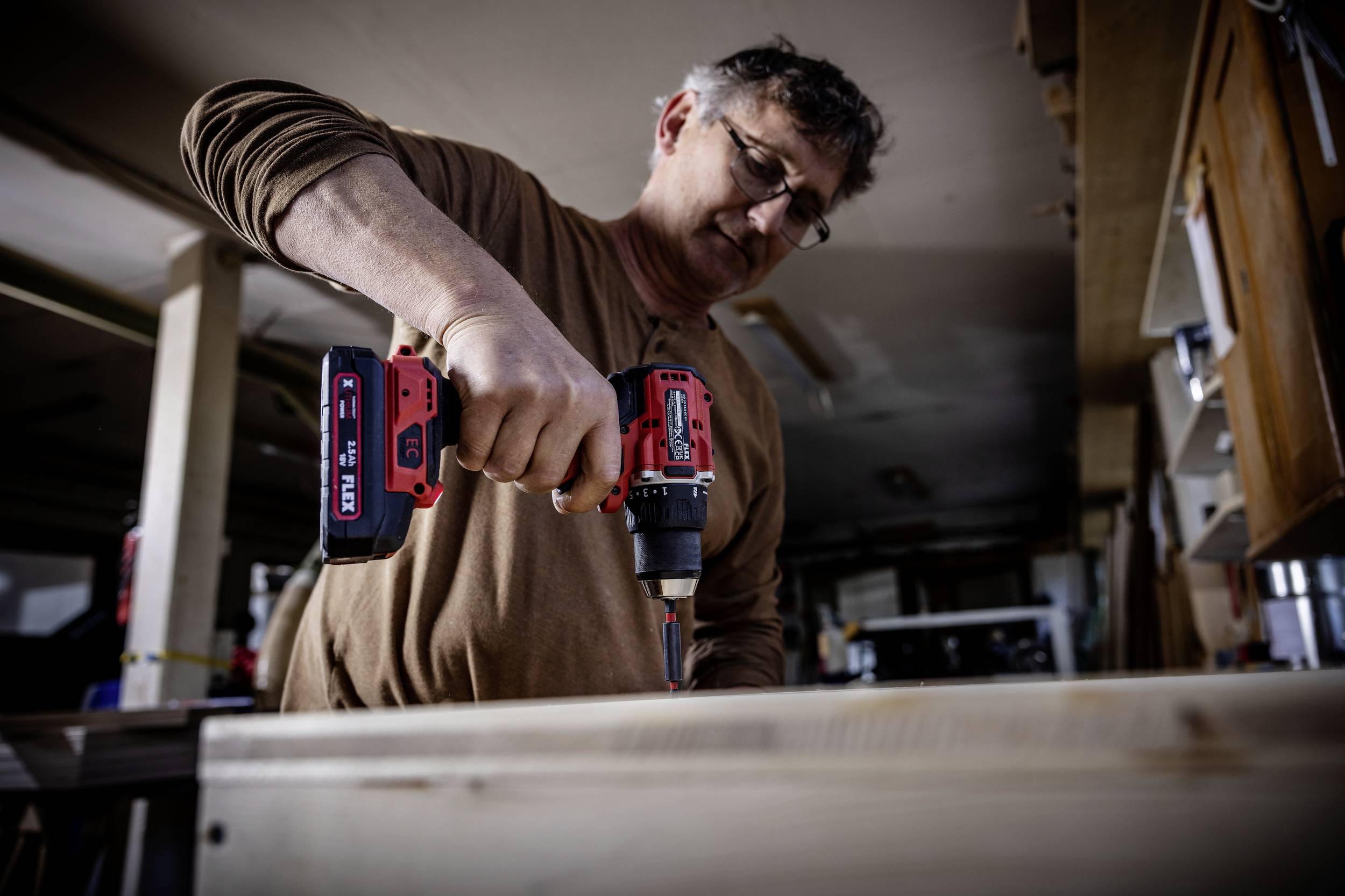 Een man werkt geconcentreerd met een accuschroevendraaier aan een houten project in een werkplaats. Op de achtergrond zijn gereedschappen zichtbaar.