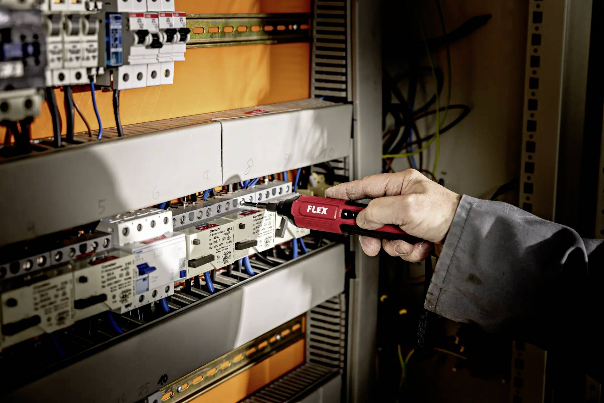 Een persoon werkt met een rode spanningzoeker aan een geopende schakelkast vol elektrische componenten en kabels.