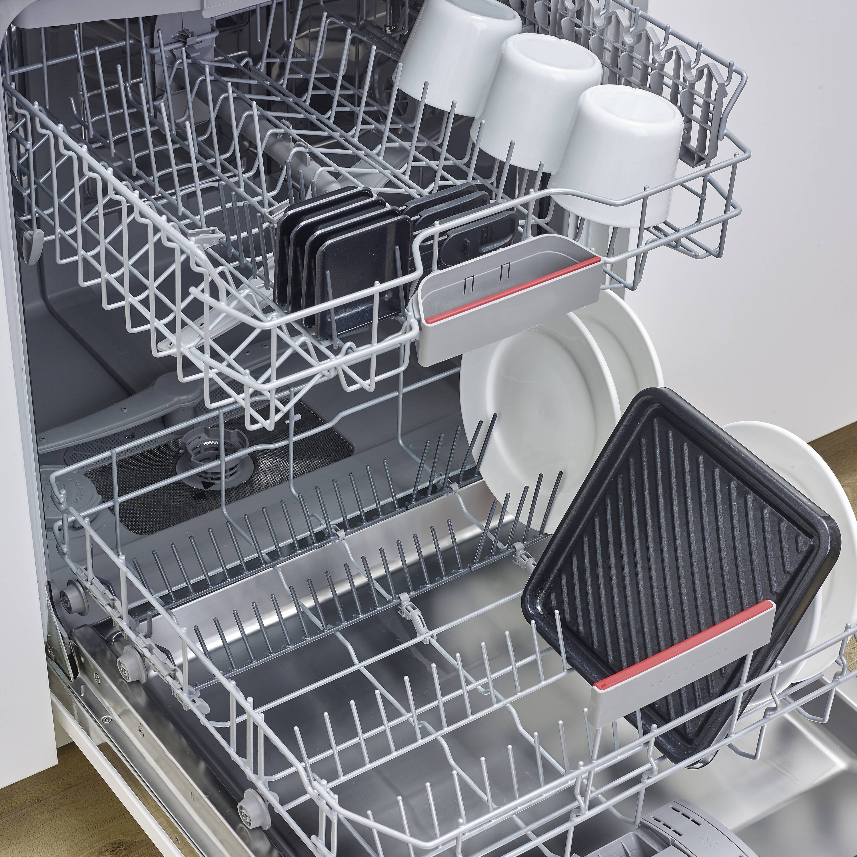 Een openstaande vaatwasser met servies: Bovenste en onderste niveau met kopjes, borden en dienbladen. Dienbladen geplaatst in bestelopbergbakken.