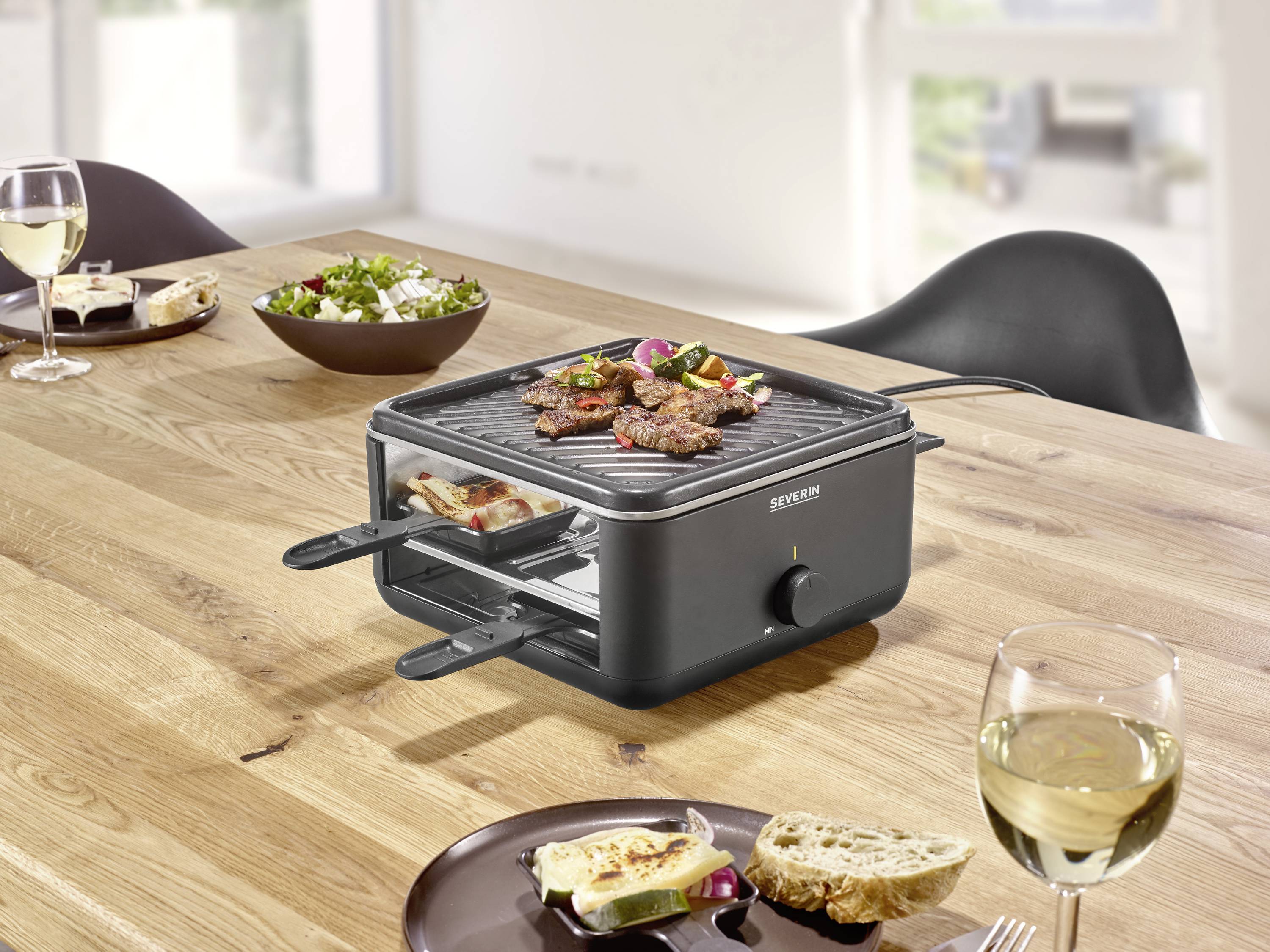 Een tafelgrill met vlees en groenten op een houten tafel met twee glazen witte wijn, salade en brood in een moderne ruimte.