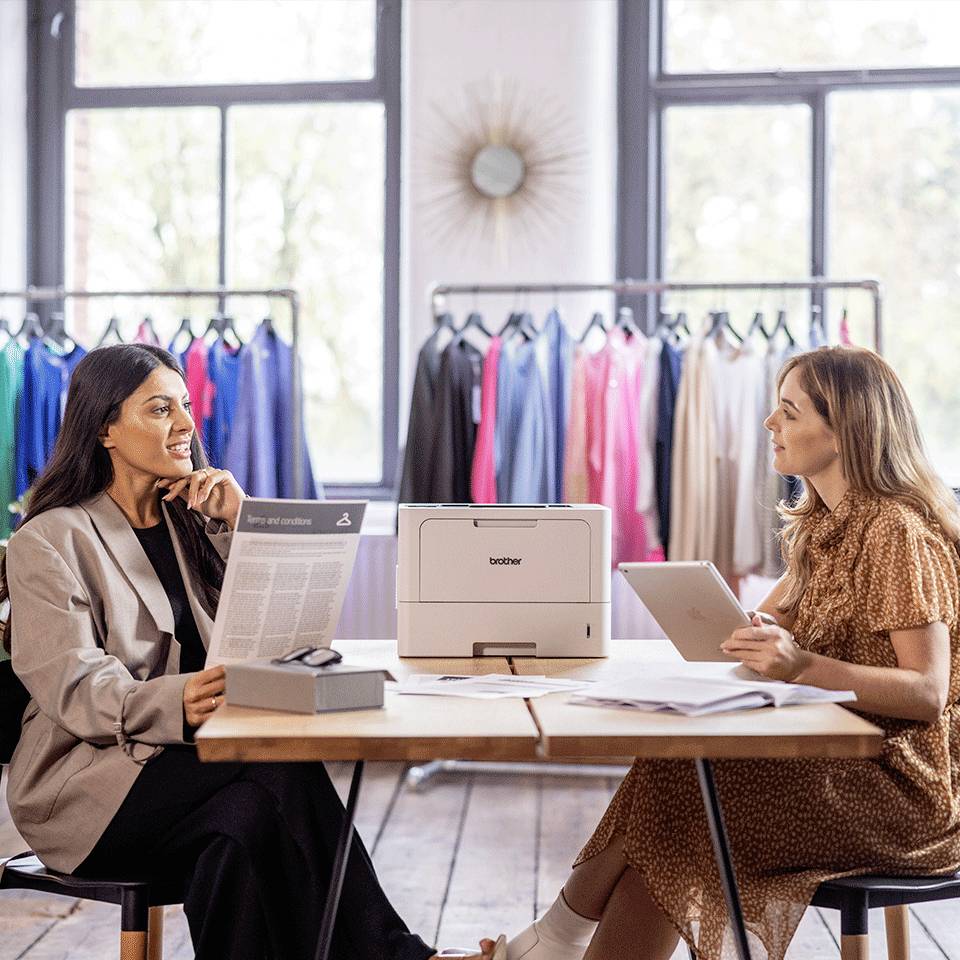 Twee vrouwen zitten aan een tafel in een modezaak, omringd door kleding. Ze bespreken documenten voor een printer.